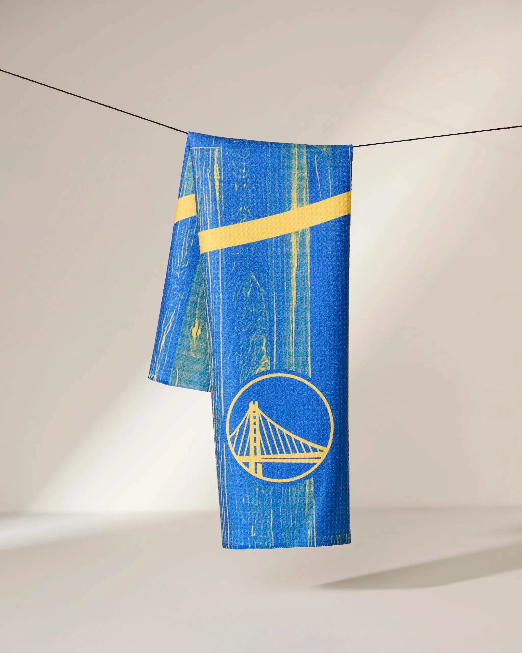 The Warriors Zone towel by Geometry, featuring a stylized bridge logo in blue and yellow, hangs over a thin black line against a plain light background.