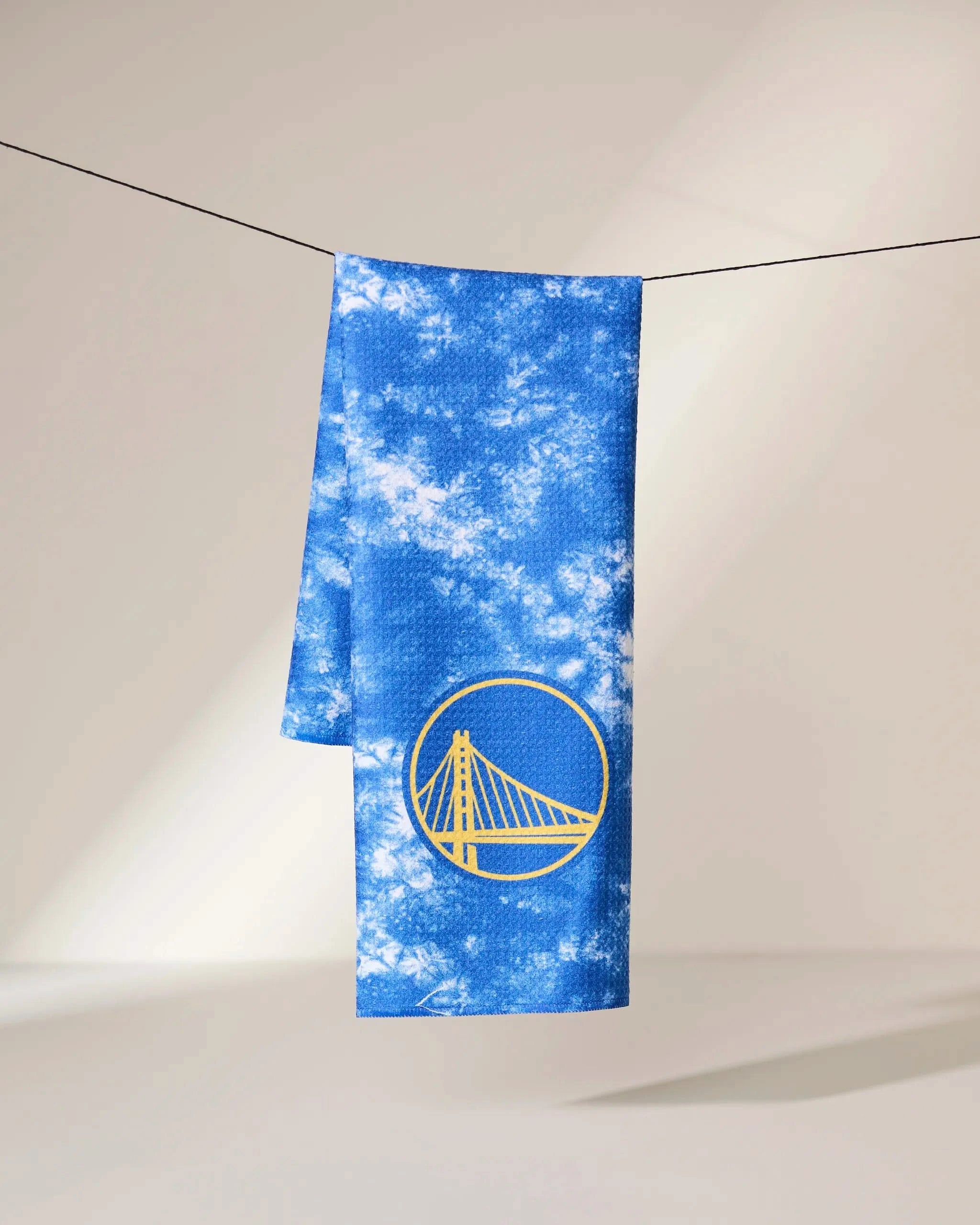 The Geometry Warriors Flash towel, featuring a blue and white tie-dye design and a yellow Golden State Warriors bridge logo, hangs on a thin black line against a light, neutral background.