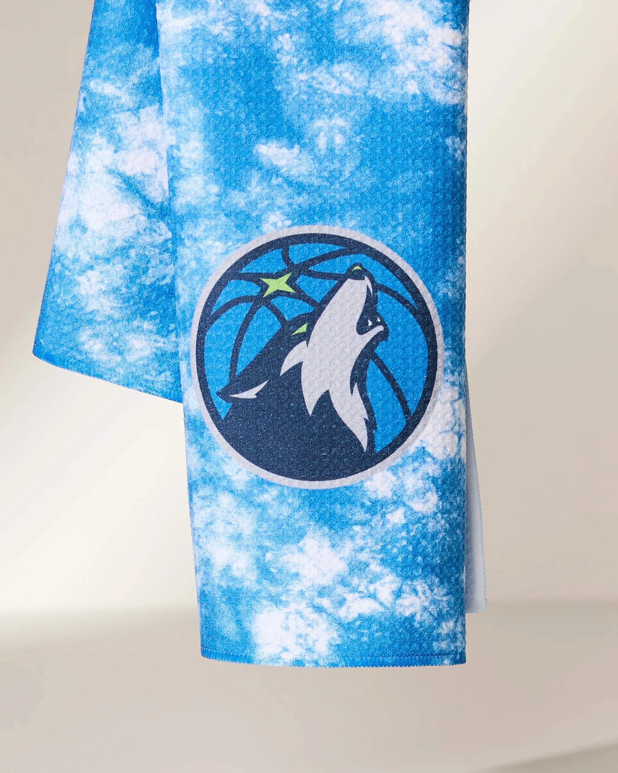 The Geometry Timberwolves Flash towel features a blue and white tie-dye design with the Minnesota Timberwolves logo—a gray wolf howling at a star in front of a basketball.