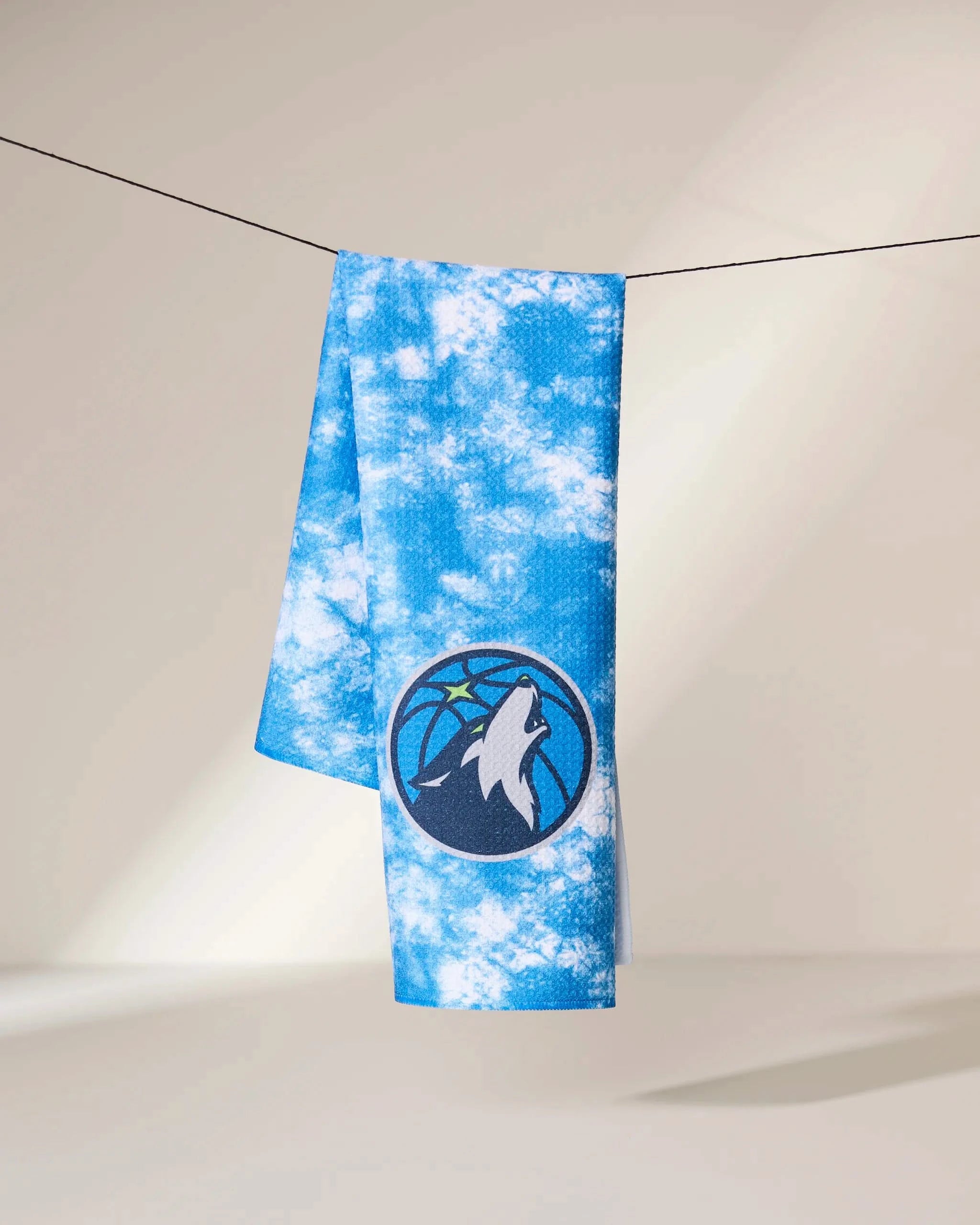 A Timberwolves Flash towel by Geometry, featuring a blue and white tie-dye design with the Minnesota Timberwolves logo, hangs over a black string against a neutral background.