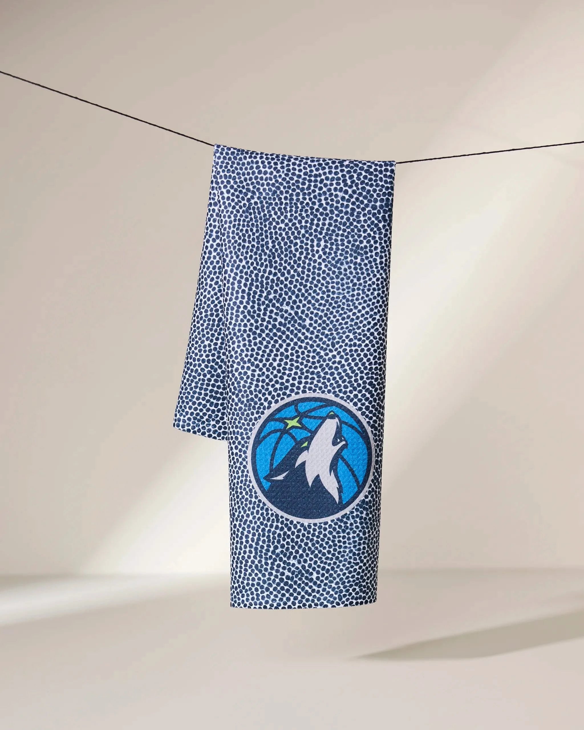 A blue and white Geometry Timberwolves Grip towel hangs on a line, featuring the Minnesota Timberwolves logo with a howling wolf, basketball, and pine trees. The softly lit background is neutral.