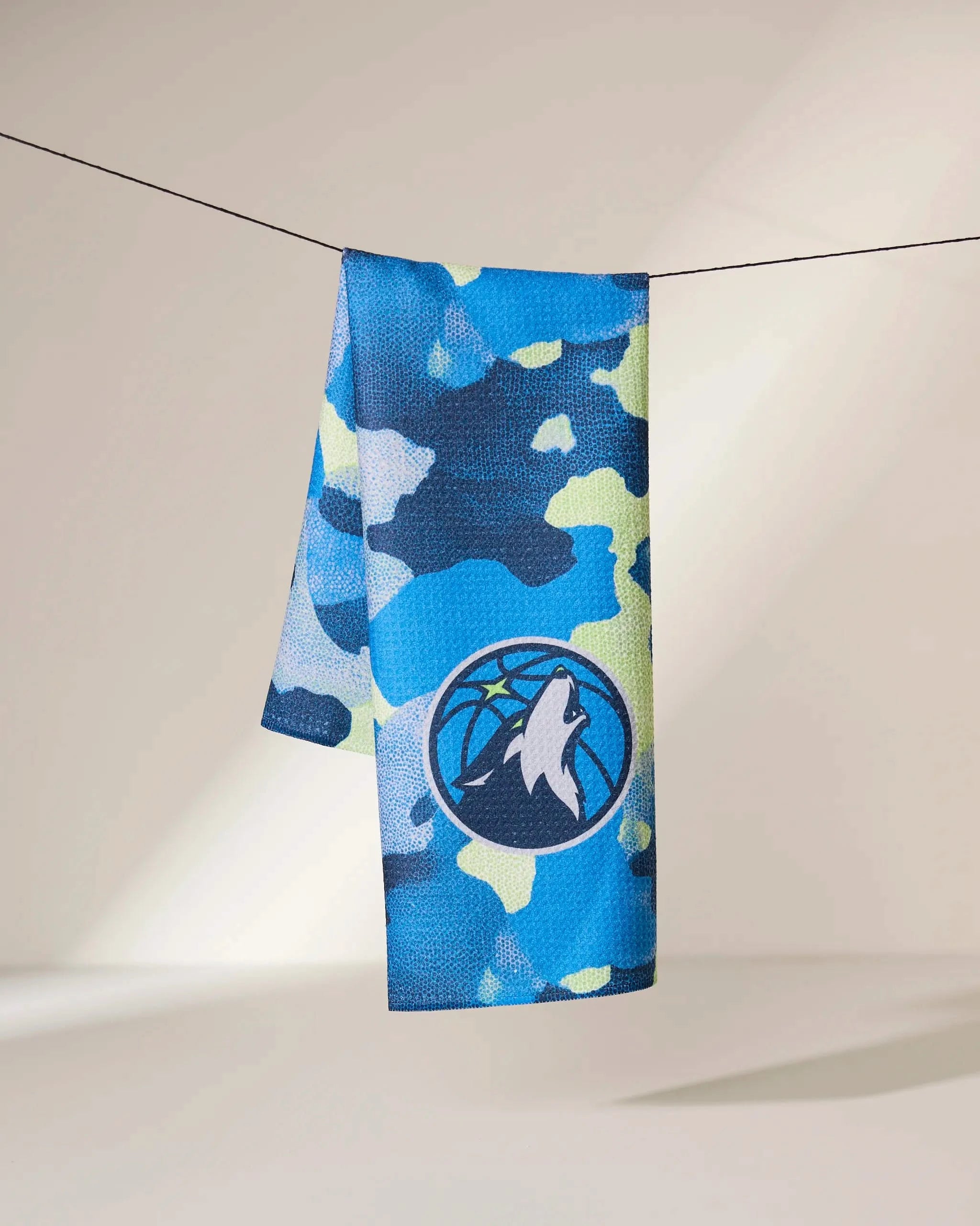 The Timberwolves Camo towel by Geometry, featuring a blue, green, and yellow camouflage pattern with a basketball wolf logo, hangs on a line against a neutral background.