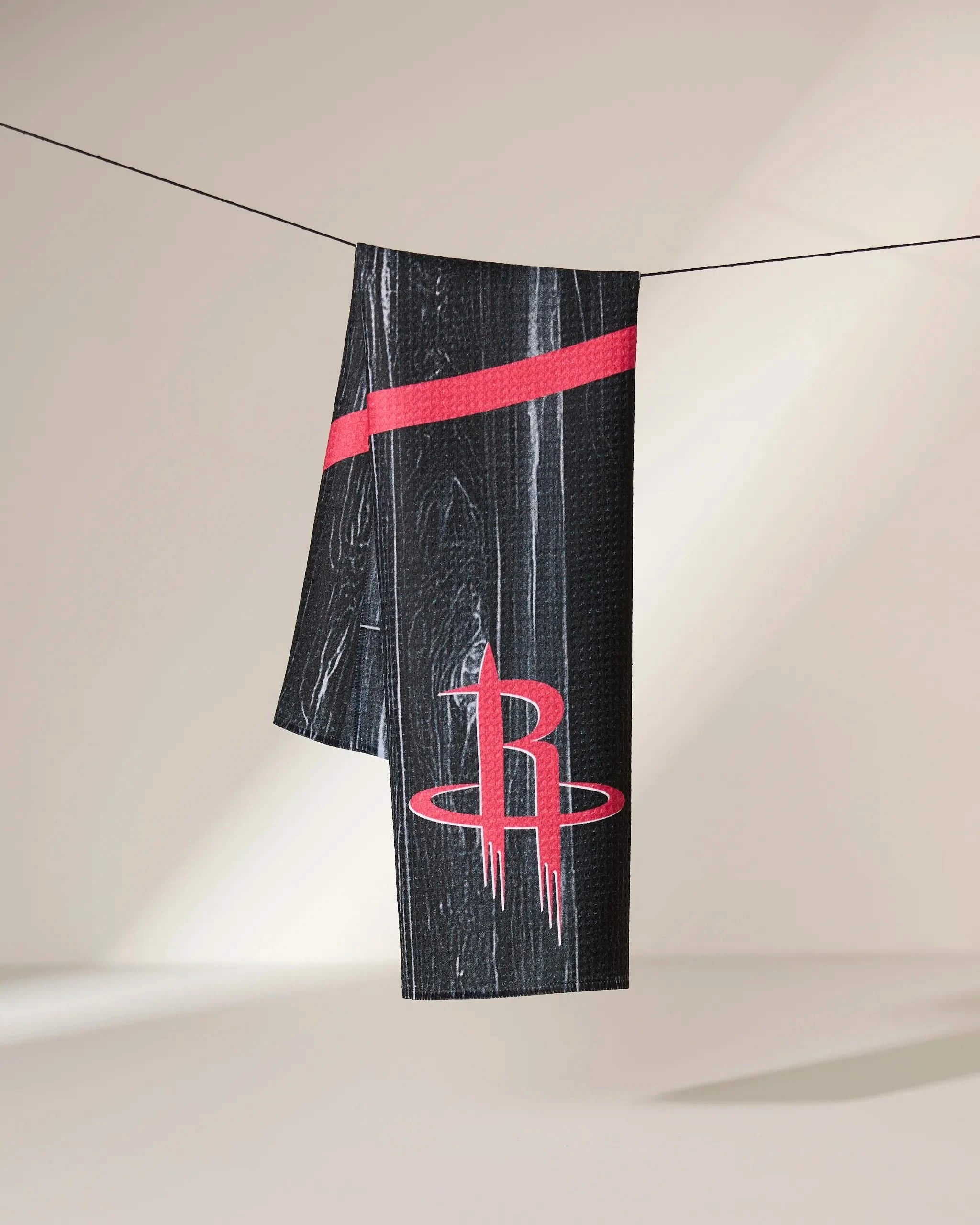 The Geometry Rockets Zone towel, featuring a black design with a red diagonal stripe and Houston Rockets logo, hangs on a string against a light, neutral background.