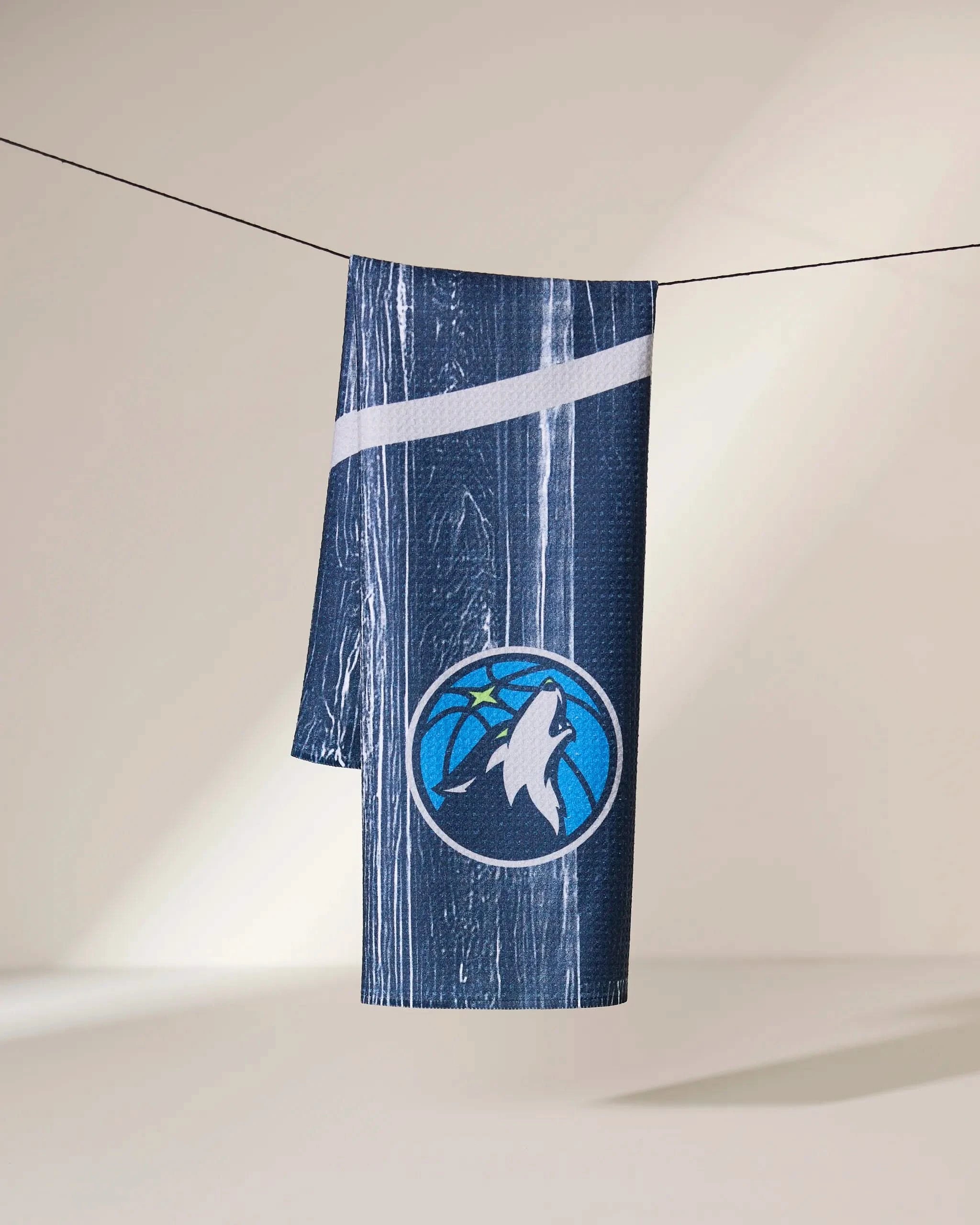 The Geometry Timberwolves Zone towel, featuring navy blue fabric with white stripes and the Minnesota Timberwolves logo, hangs on a clothesline against a neutral background.