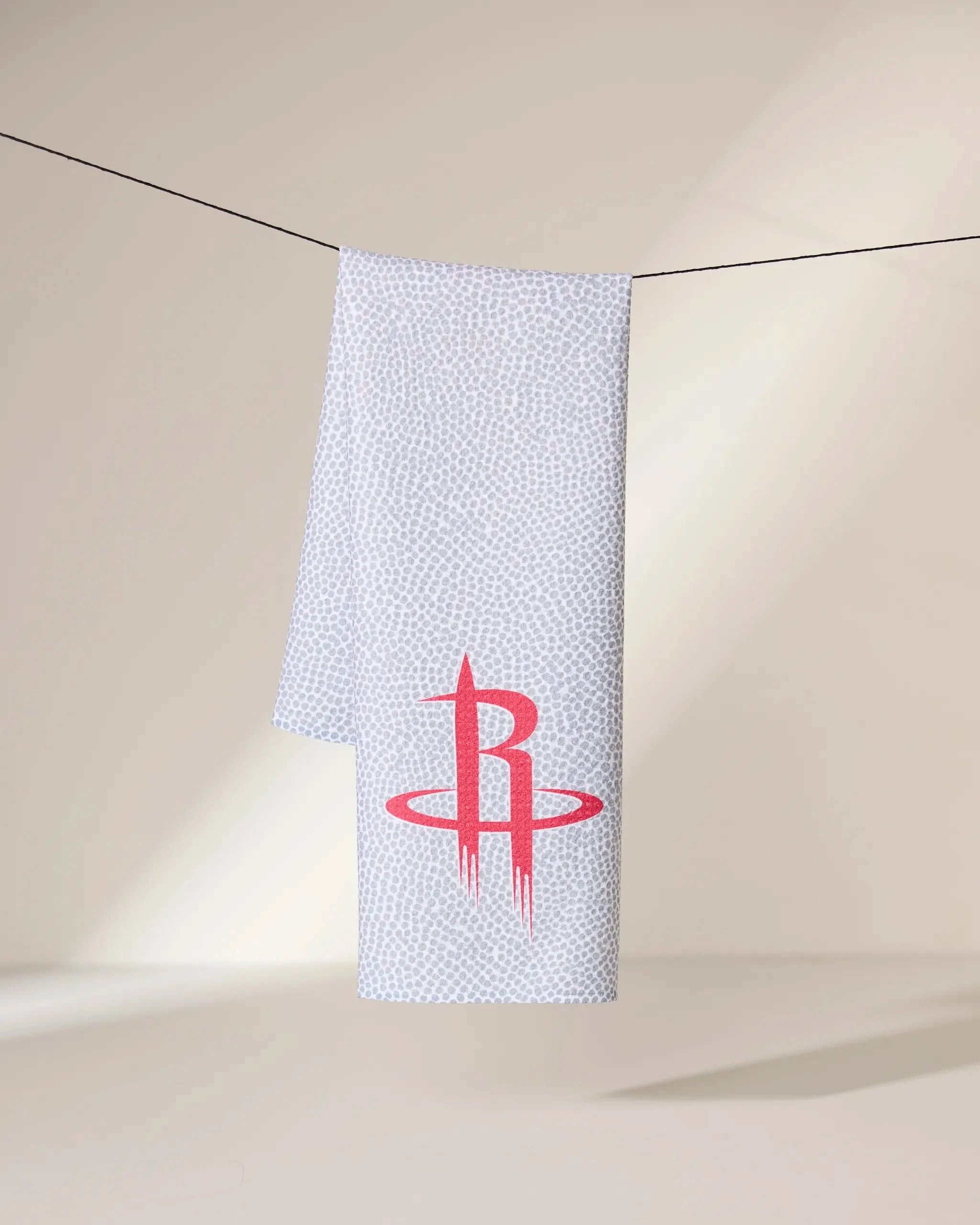 A gray Geometry Rockets Grip towel with a subtle dotted pattern hangs on a black line, showcasing a large red Houston Rockets logo near the bottom against a neutral, softly lit background.