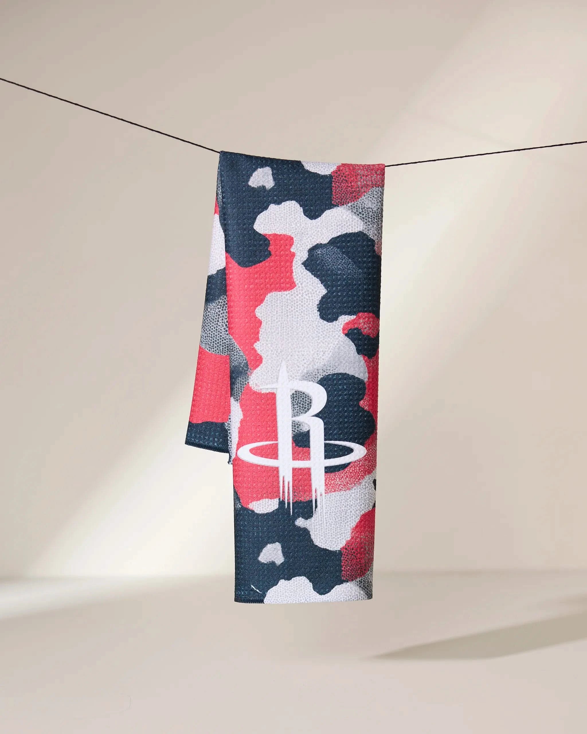 The Geometry Rockets Camo towel, featuring a red, black, white, and gray camouflage pattern with a bold white R logo inspired by the Houston Rockets, hangs on a line.