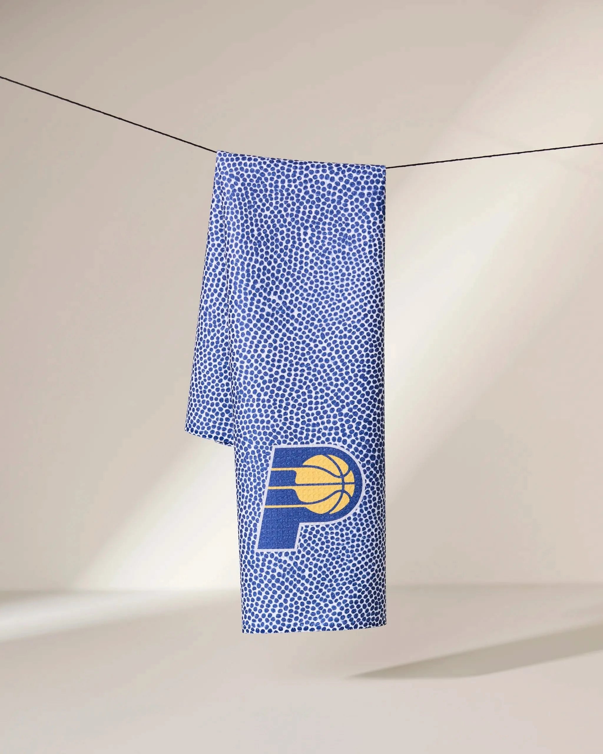 A Pacers Grip towel by Geometry, featuring a white dotted pattern and Indiana Pacers basketball logo, hangs on a clothesline against a light background.