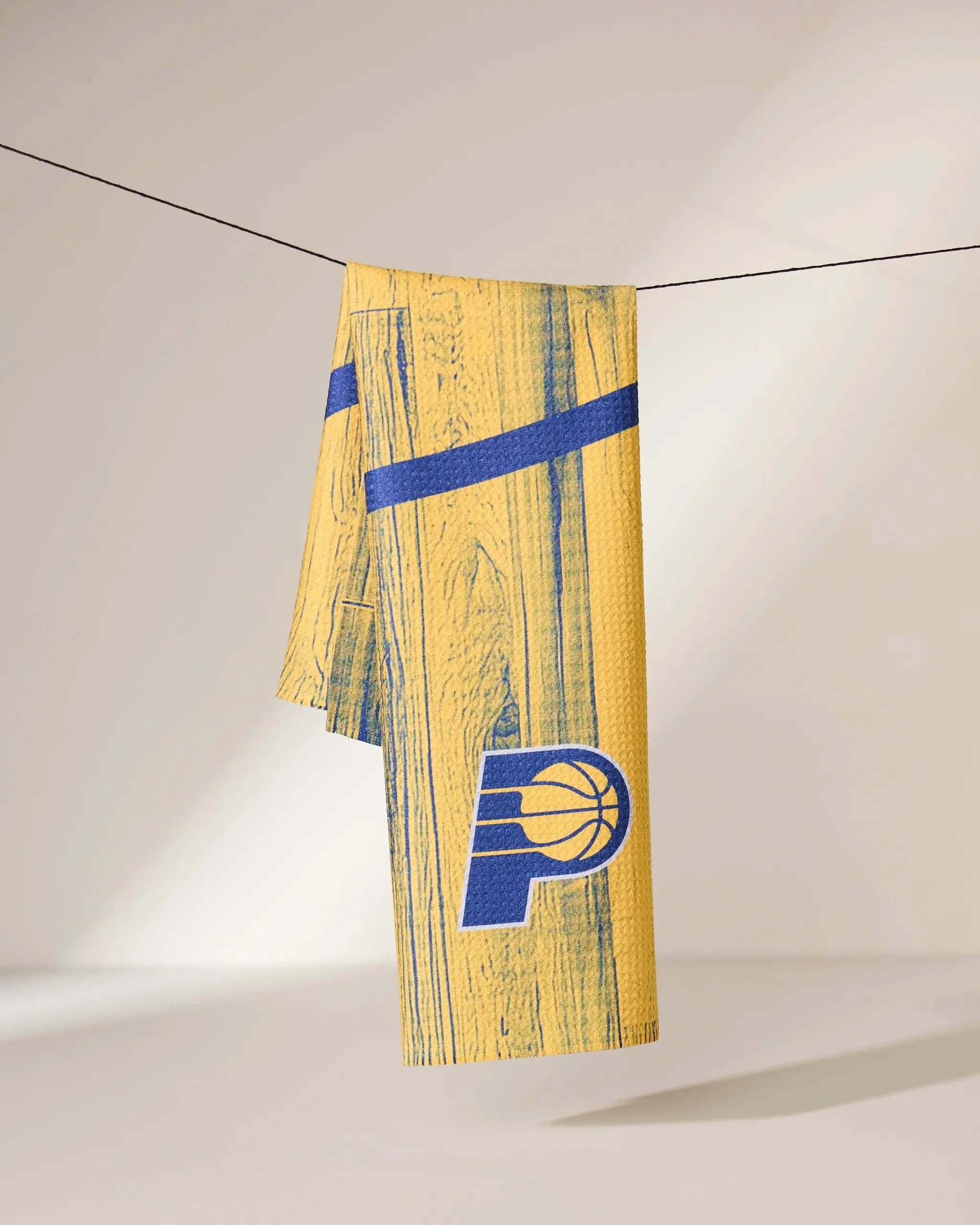 The Geometry Pacers Zone towel, featuring a yellow and blue woodgrain design with the Indiana Pacers logo, hangs on a clothesline in a bright, minimal space.