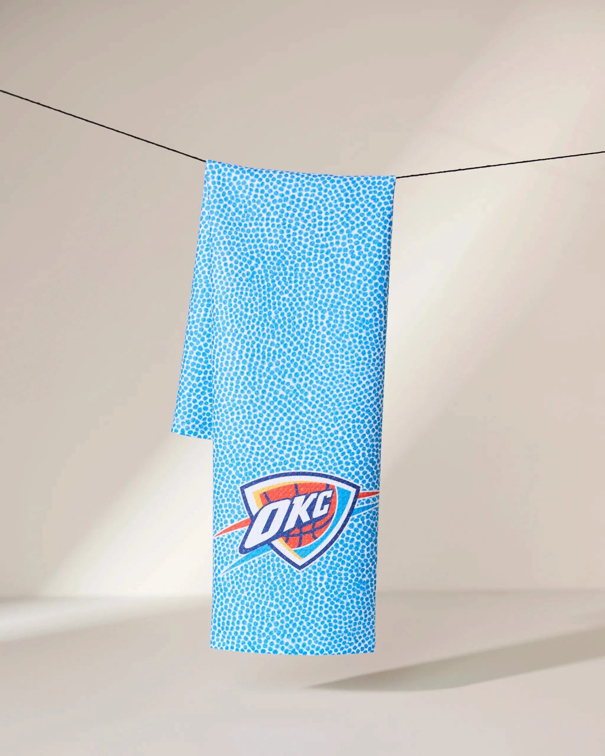 A Geometry OKC Grip towel in blue with a dotted pattern and the Oklahoma City Thunder logo hangs on a clothesline against a neutral background.