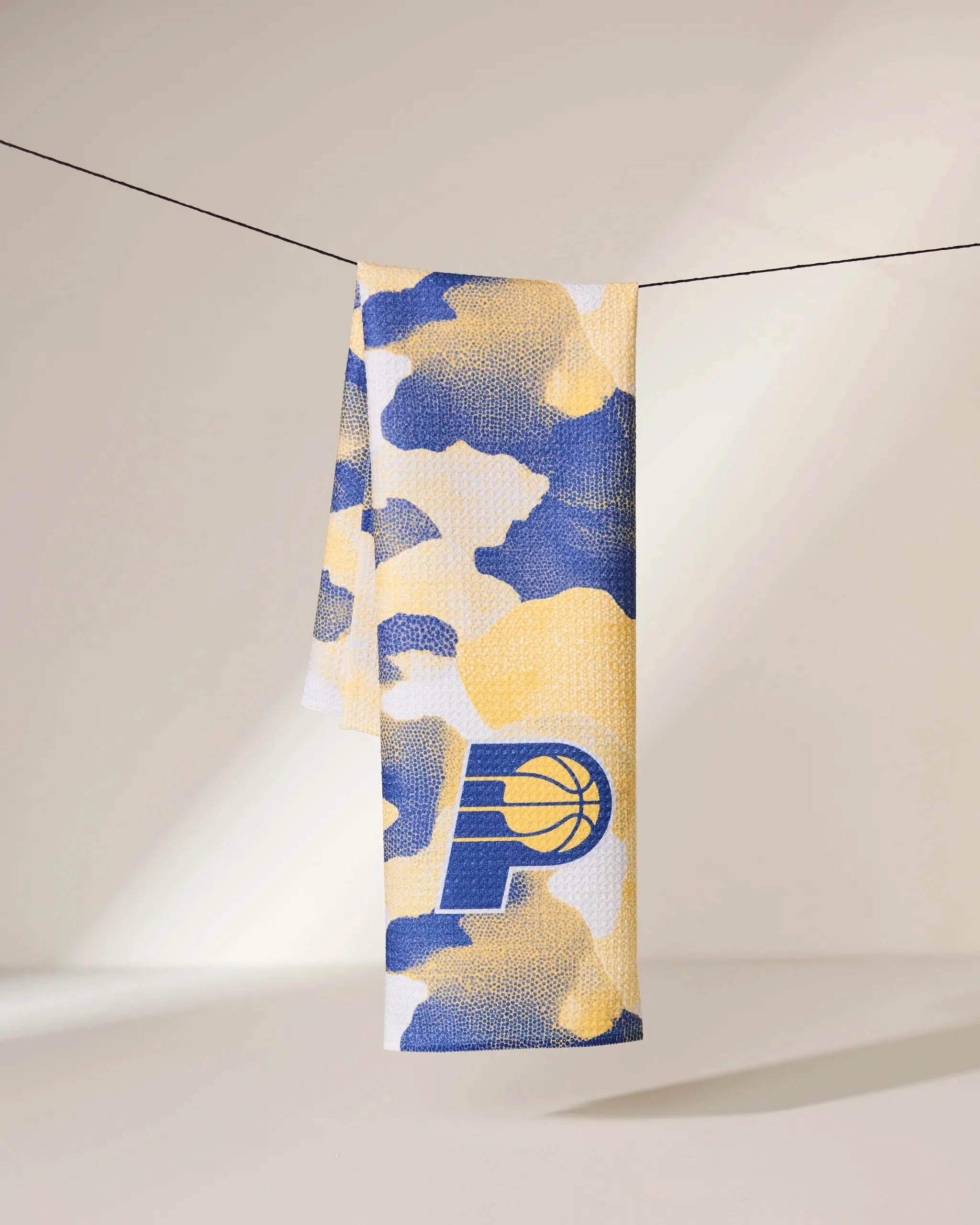 The Geometry Pacers Camo towel, featuring a blue, yellow, and white camouflage pattern and the Indiana Pacers logo, hangs on a clothesline against a softly lit background.