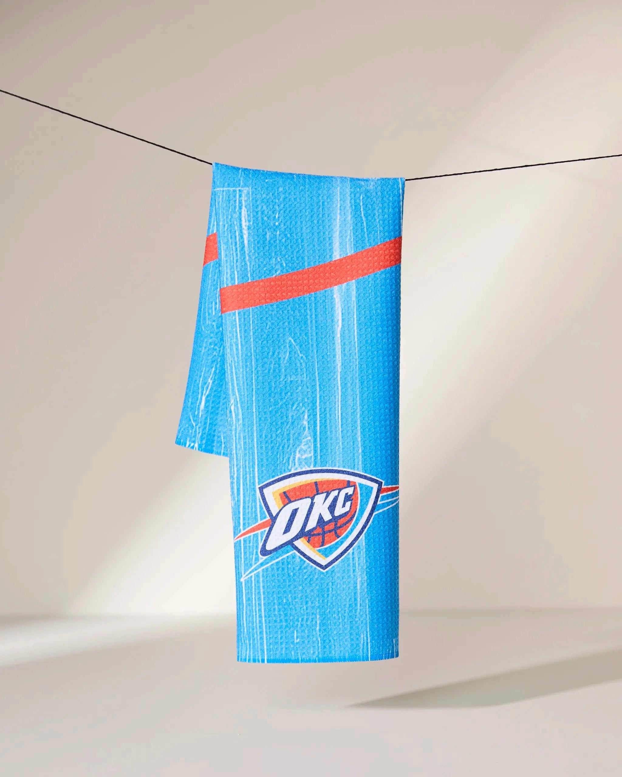 The OKC Zone towel by Geometry, featuring orange stripes and the Oklahoma City Thunder logo, hangs over a black clothesline against a neutral background.