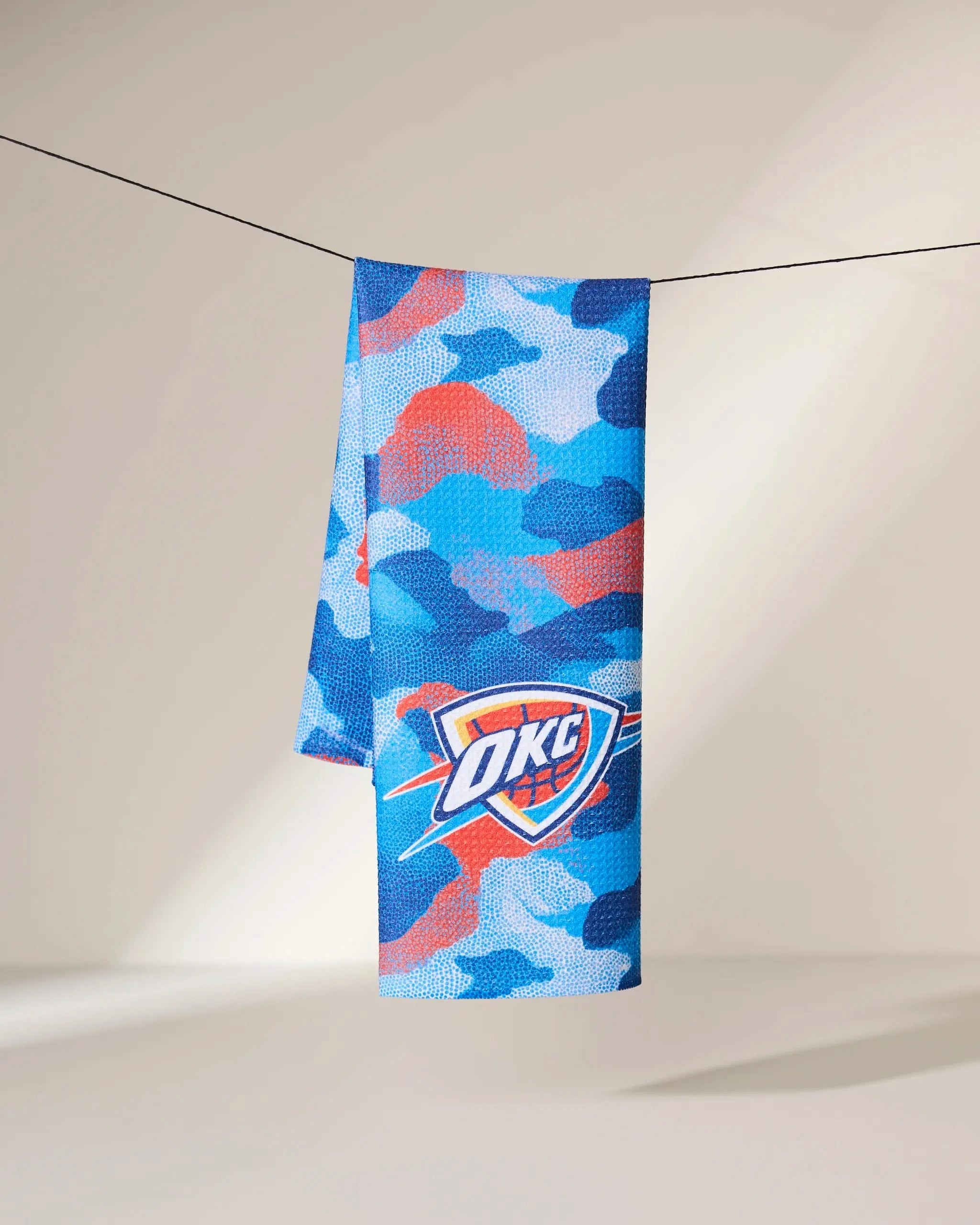 The Geometry OKC Camo towel, featuring a blue, red, and white camouflage pattern with the OKC Thunder logo, hangs on a black line against a neutral background.