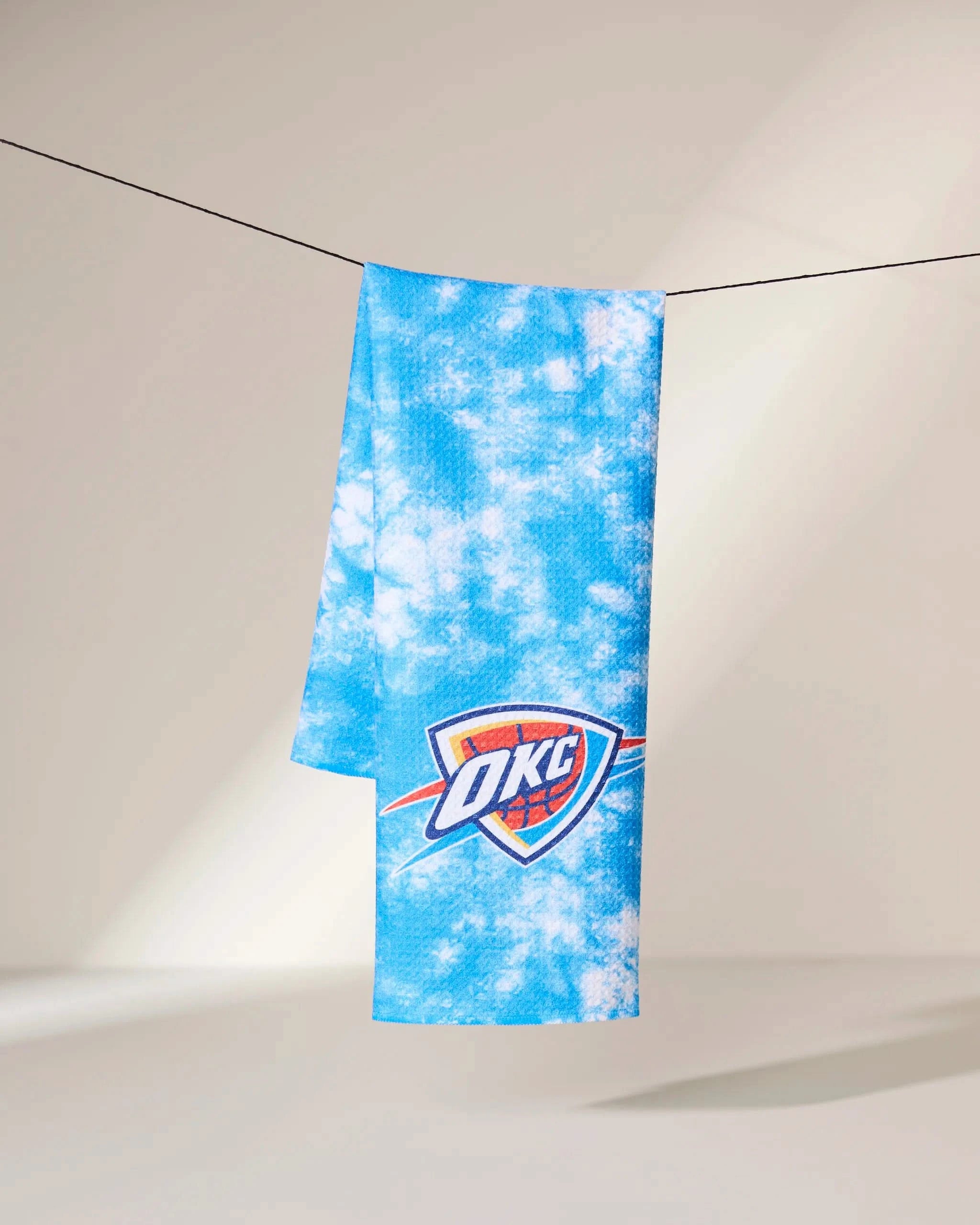 The Geometry OKC Flash towel, featuring a blue and white tie-dye design and the Oklahoma City Thunder logo, hangs from a black string against a plain light background.