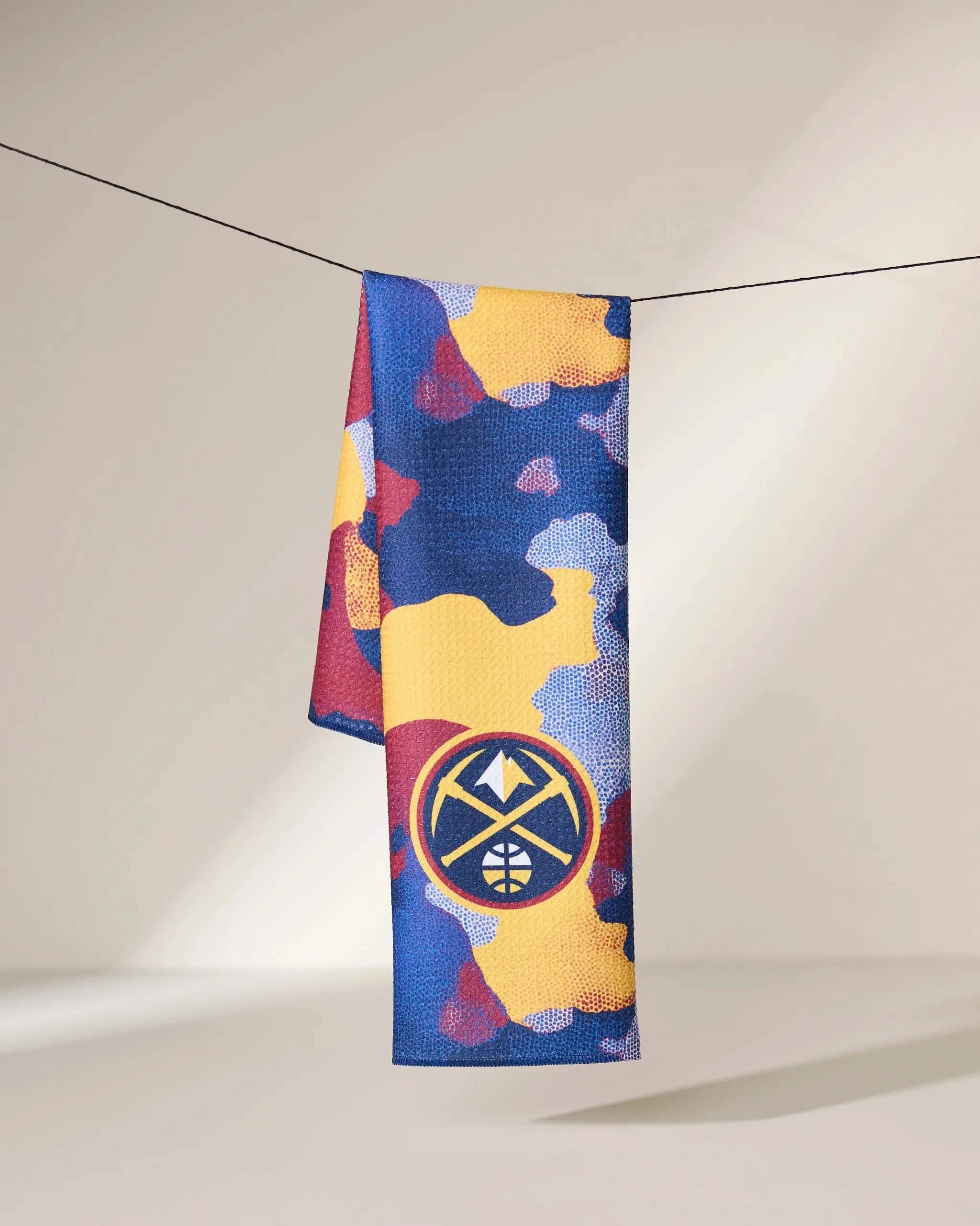 The Geometry Nuggets Camo towel displays blue, yellow, and red abstract patterns with a white and gold stylized basketball and mountain logo near the bottom, hanging on a line.