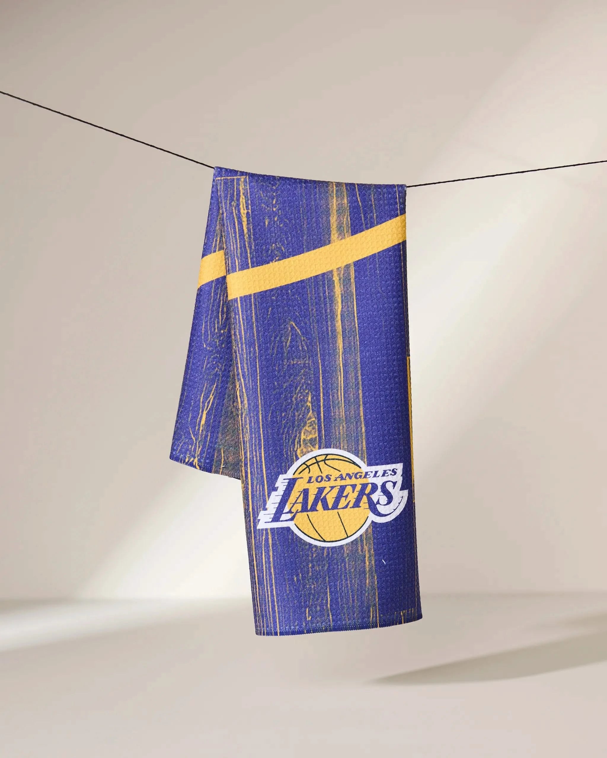 A purple and gold Geometry Lakers Zone towel with the Los Angeles Lakers logo, wood grain textures, and diagonal yellow stripes hangs on a clothesline against a softly lit plain background.