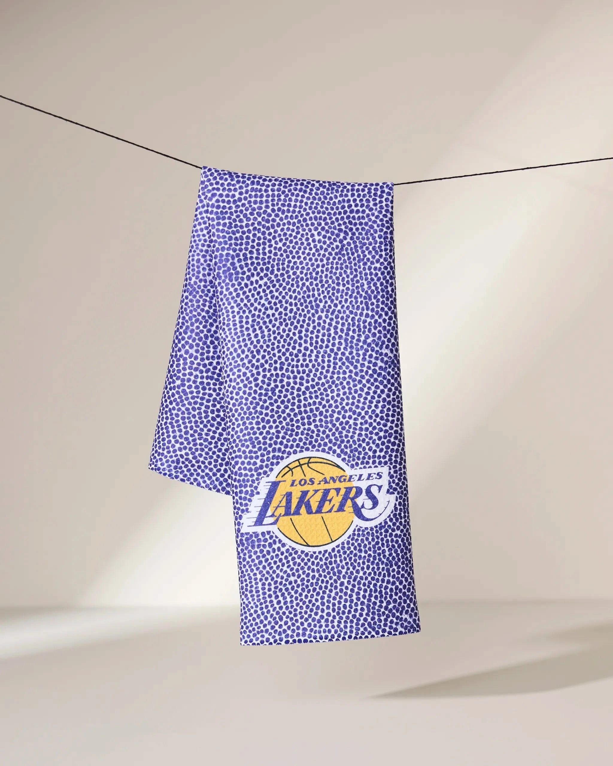 A purple Geometry Lakers Grip towel with a textured dot pattern hangs on a line, featuring the Los Angeles Lakers logo in bold purple and gold lettering over a yellow basketball graphic.