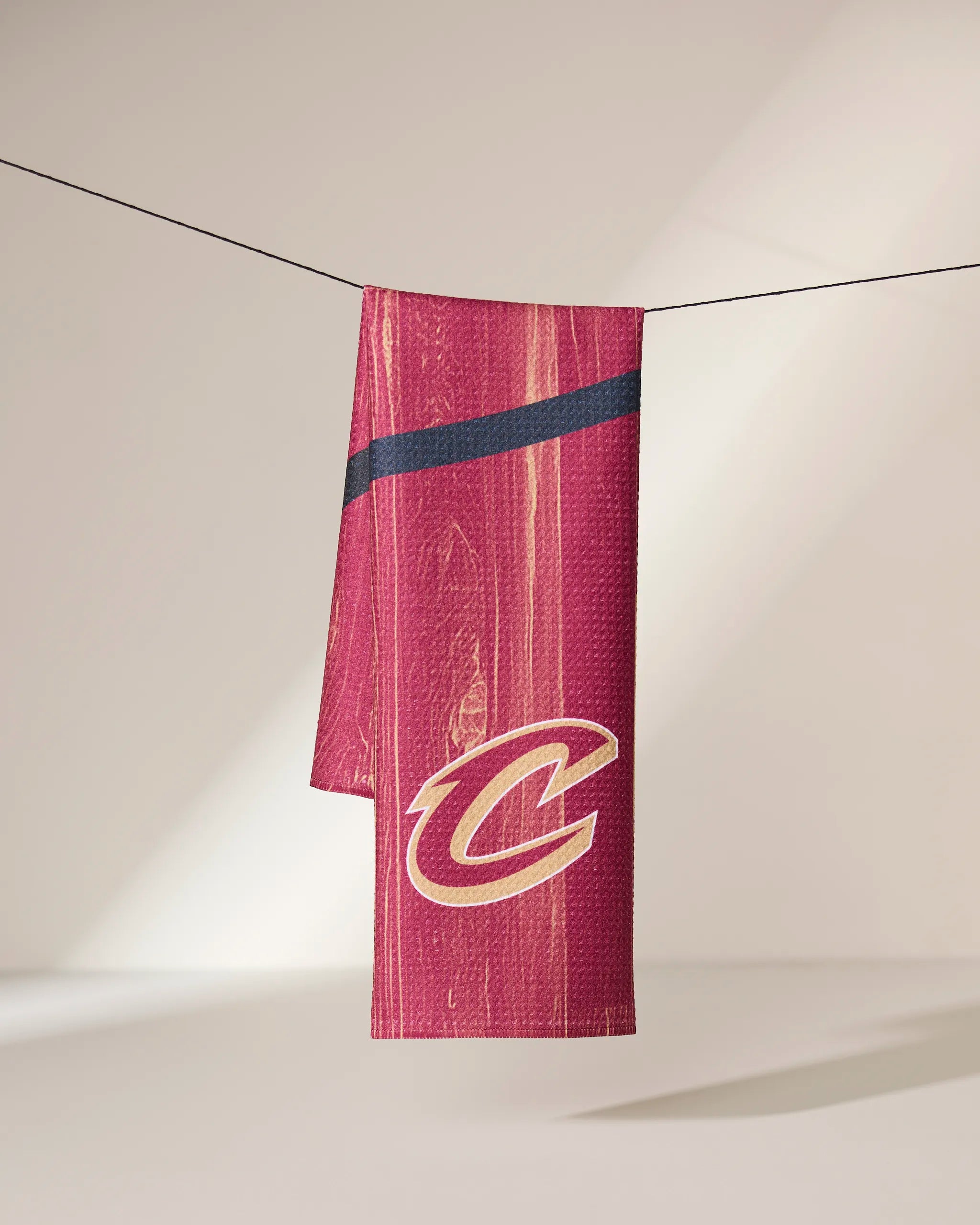 The Geometry Cavaliers Zone towel, featuring a maroon color, yellow Cleveland Cavaliers C logo, and dark diagonal stripe, hangs on a thin line against a softly lit background.