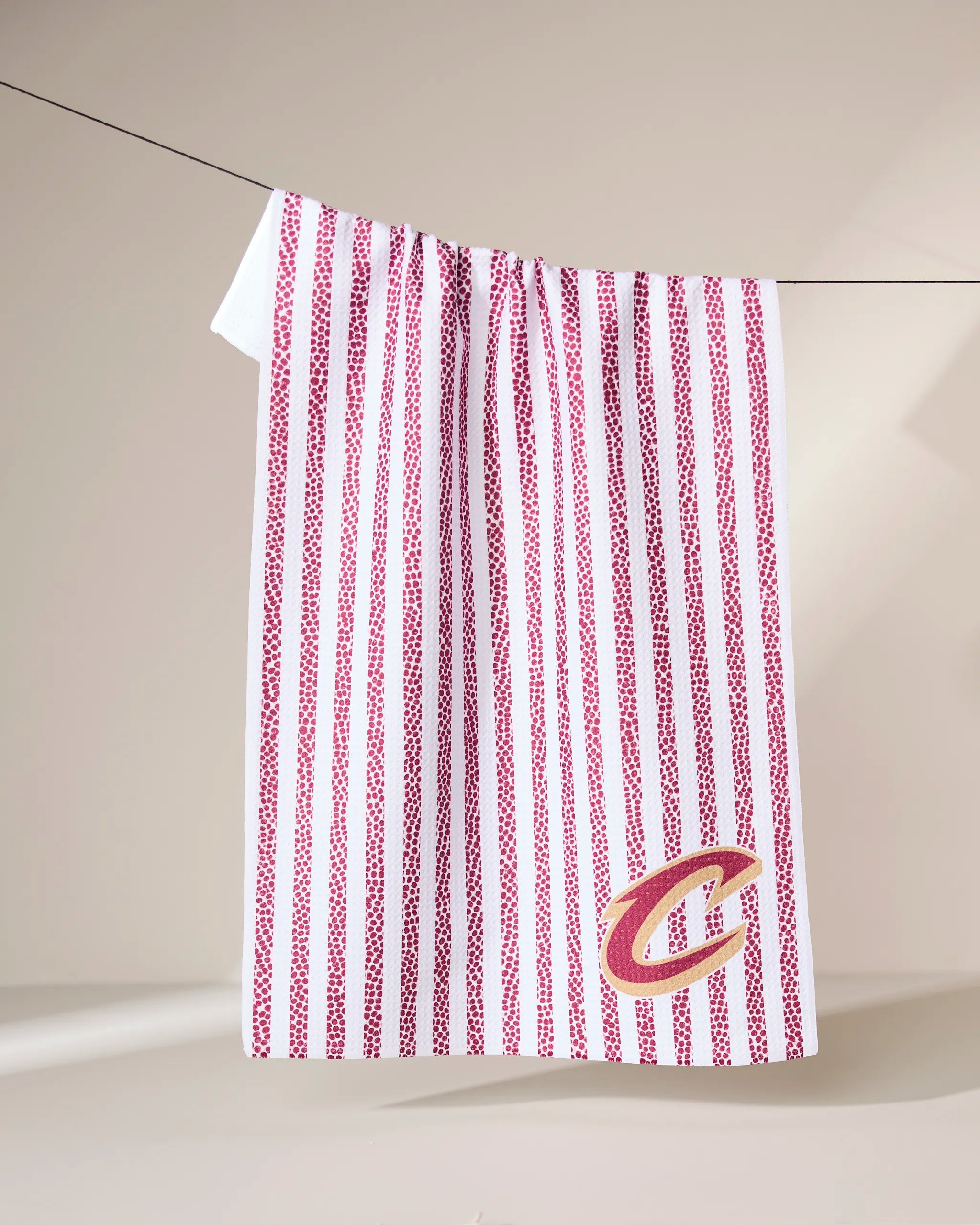 The Geometry Cavaliers Grip Stripe towel, white with vertical red stripes, hangs on a line. A stylized gold and red C is embroidered near the bottom right corner against a neutral, softly lit background.