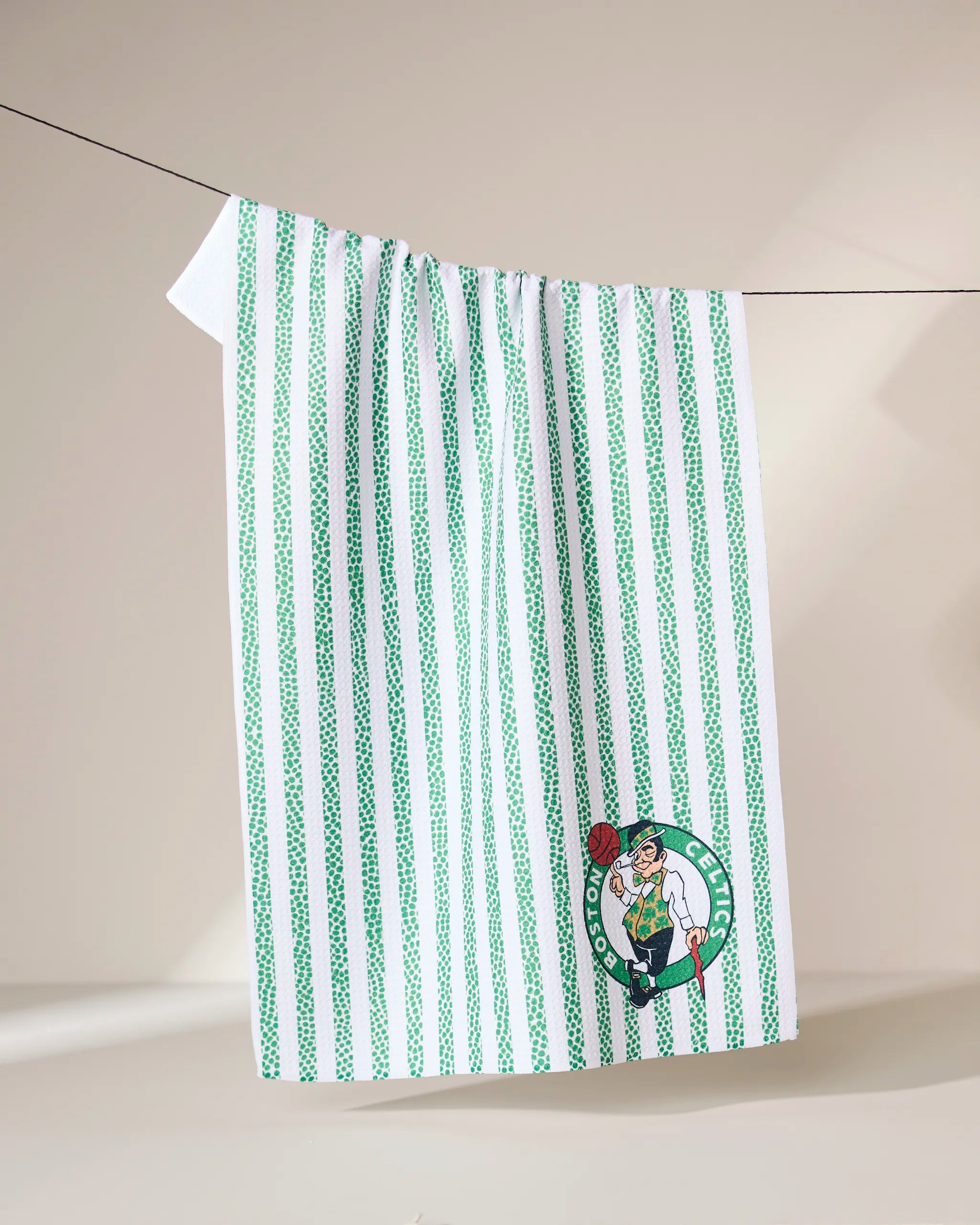 The Geometry Celtics Grip Stripe towel, featuring vertical green stripes and the Boston Celtics logo, hangs on a clothesline against a neutral background.