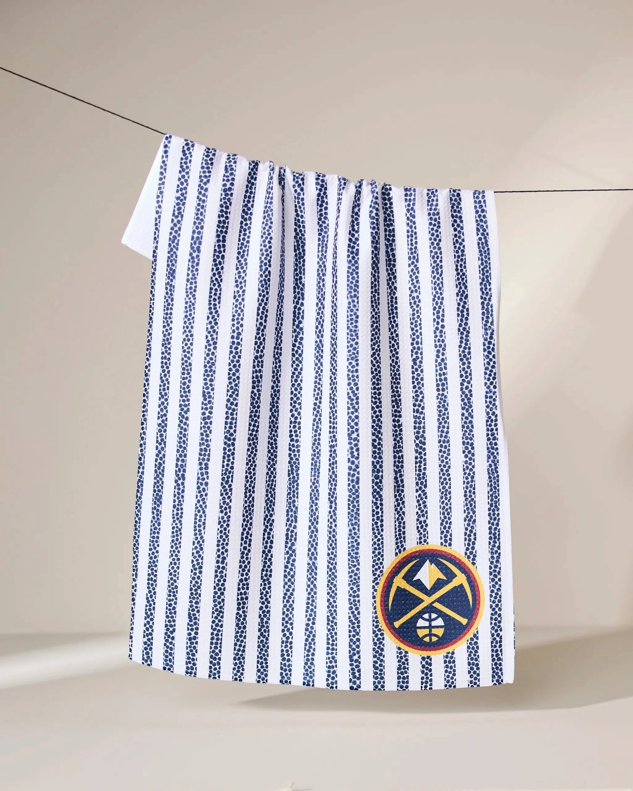 The Geometry Nuggets Grip Stripe towel, white with vertical blue stripes, hangs on a line and features a circular patch in the corner with a yellow border and a blue-and-white mountain and basketball design.