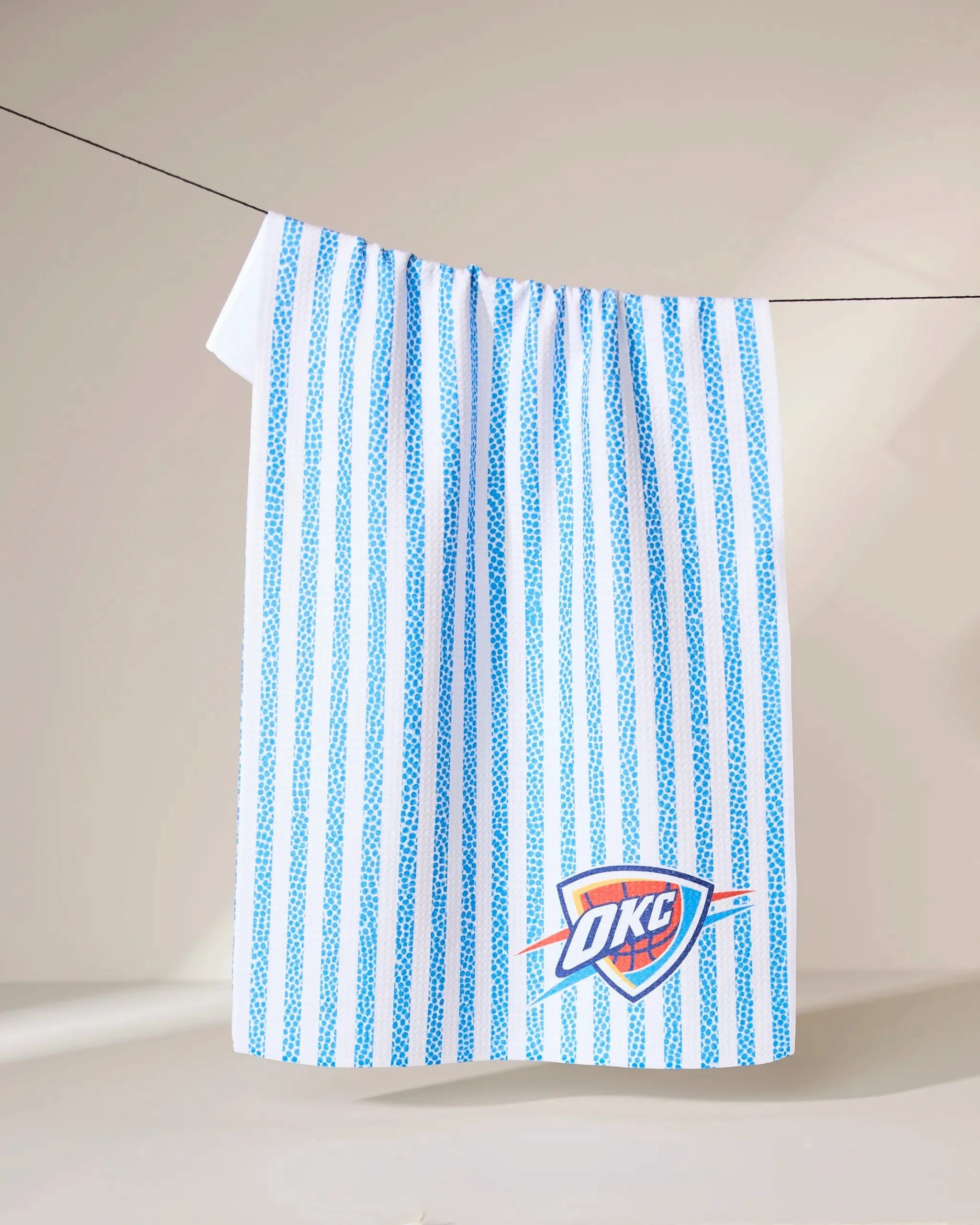 The Geometry OKC Grip Stripe towel features vertical blue stripes and the OKC Thunder logo, hanging on a clothesline against a neutral background.