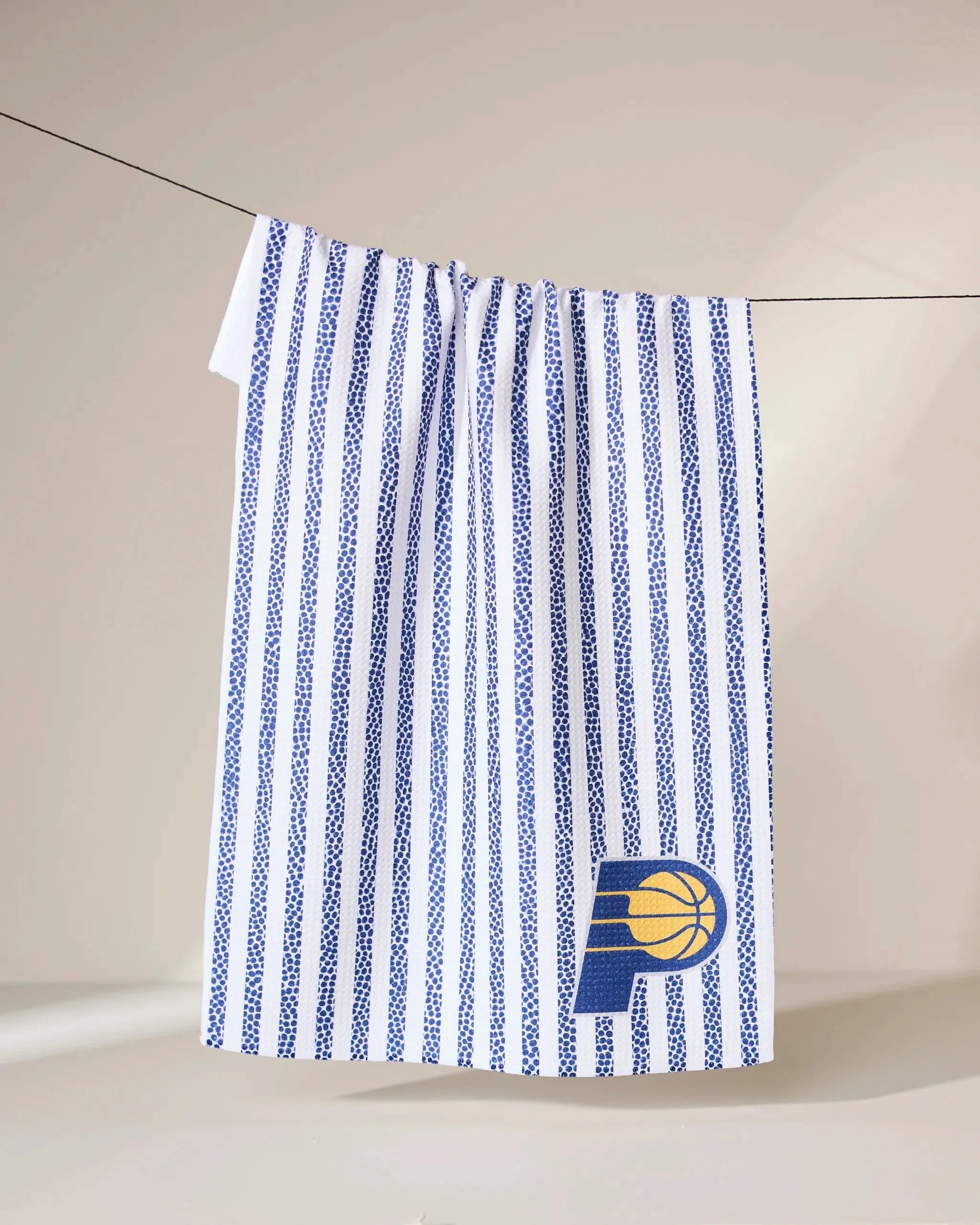 A Geometry Pacers Grip Stripe towel with vertical blue stripes hangs on a line, featuring a stylized blue P with a yellow basketball logo in the bottom right corner.