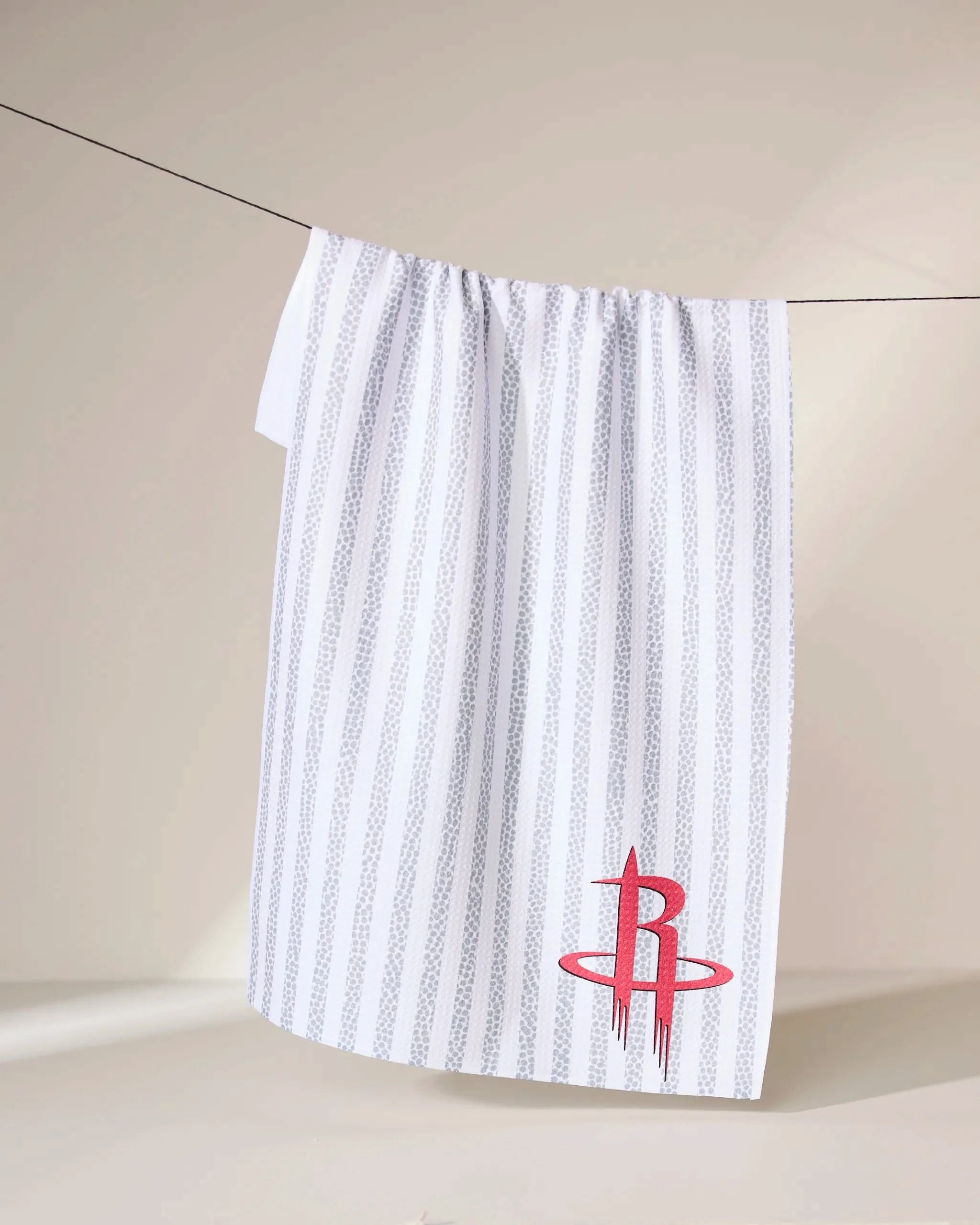 The Geometry Rockets Grip Stripe towel, featuring thin gray stripes and a red Houston Rockets logo in the bottom right corner, hangs against a plain background.