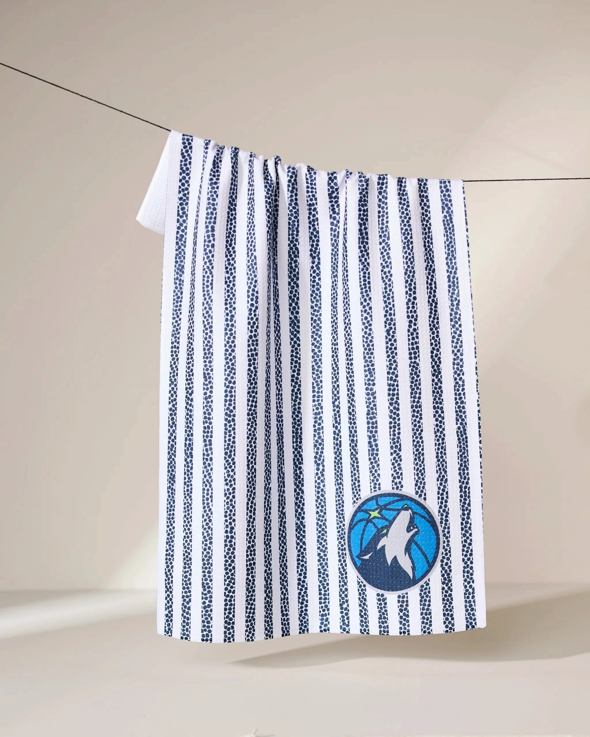 The Geometry Timberwolves Grip Stripe towel, featuring vertical navy blue stripes and a wolf logo, hangs on a clothesline against a light beige background.