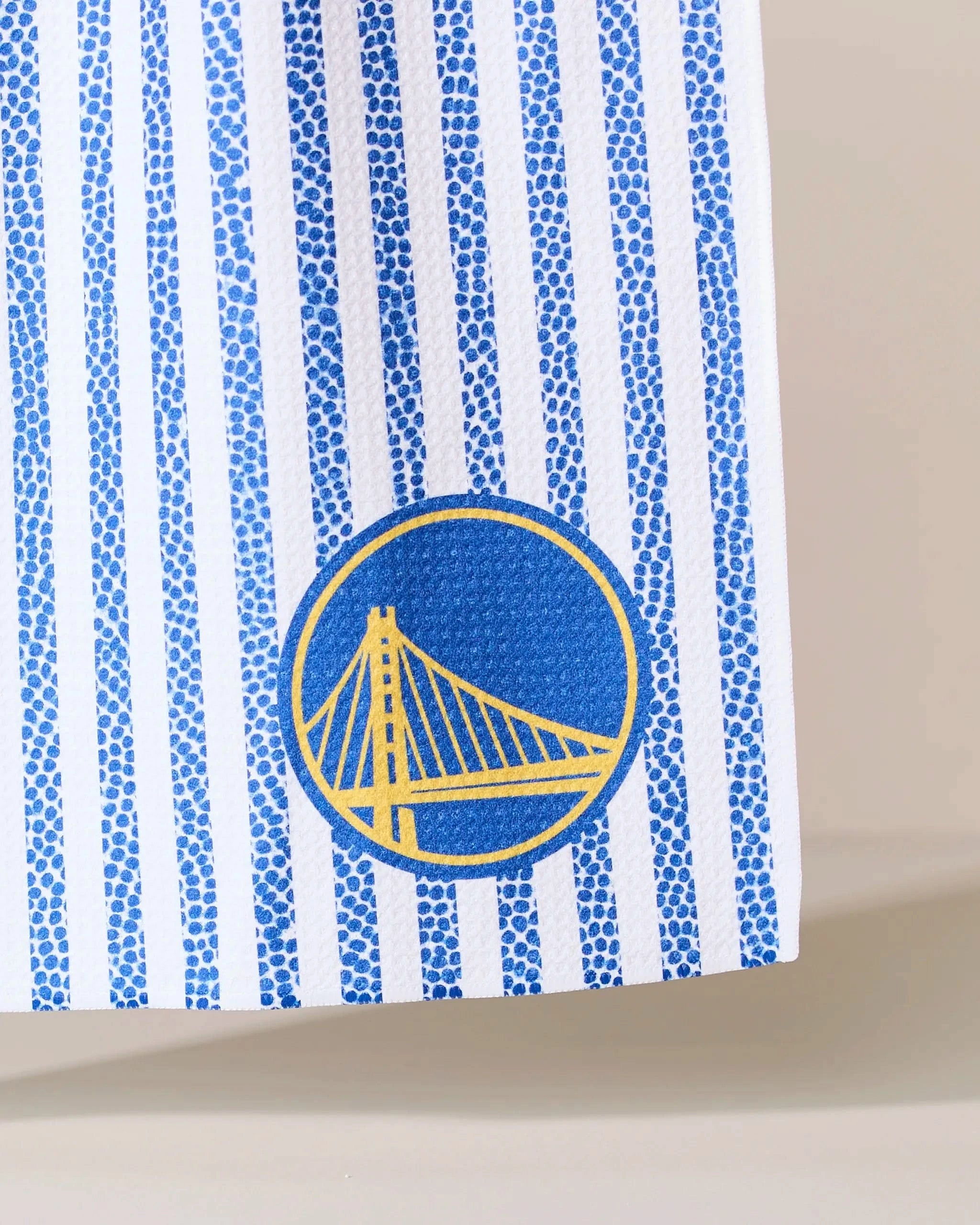 A close-up of Geometry’s Warriors Grip Stripe: a white surface with vertical blue dotted stripes and a blue circle outlined in yellow featuring a suspension bridge, inspired by the Golden State Warriors logo.