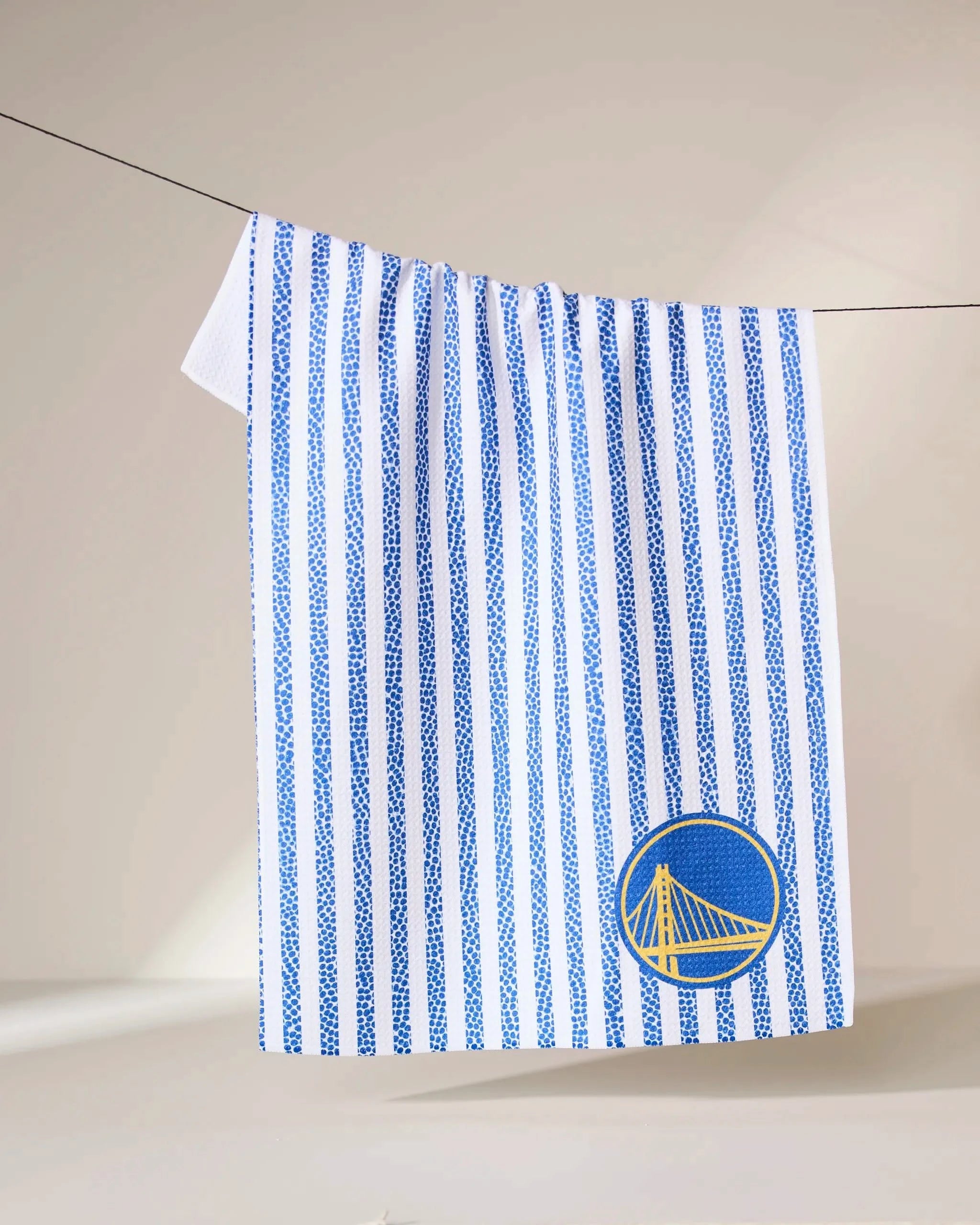 The Geometry Warriors Grip Stripe towel features vertical blue stripes and a Golden State Warriors-inspired blue and yellow circular logo in the lower right corner. It hangs on a clothesline against a plain, light-colored background.