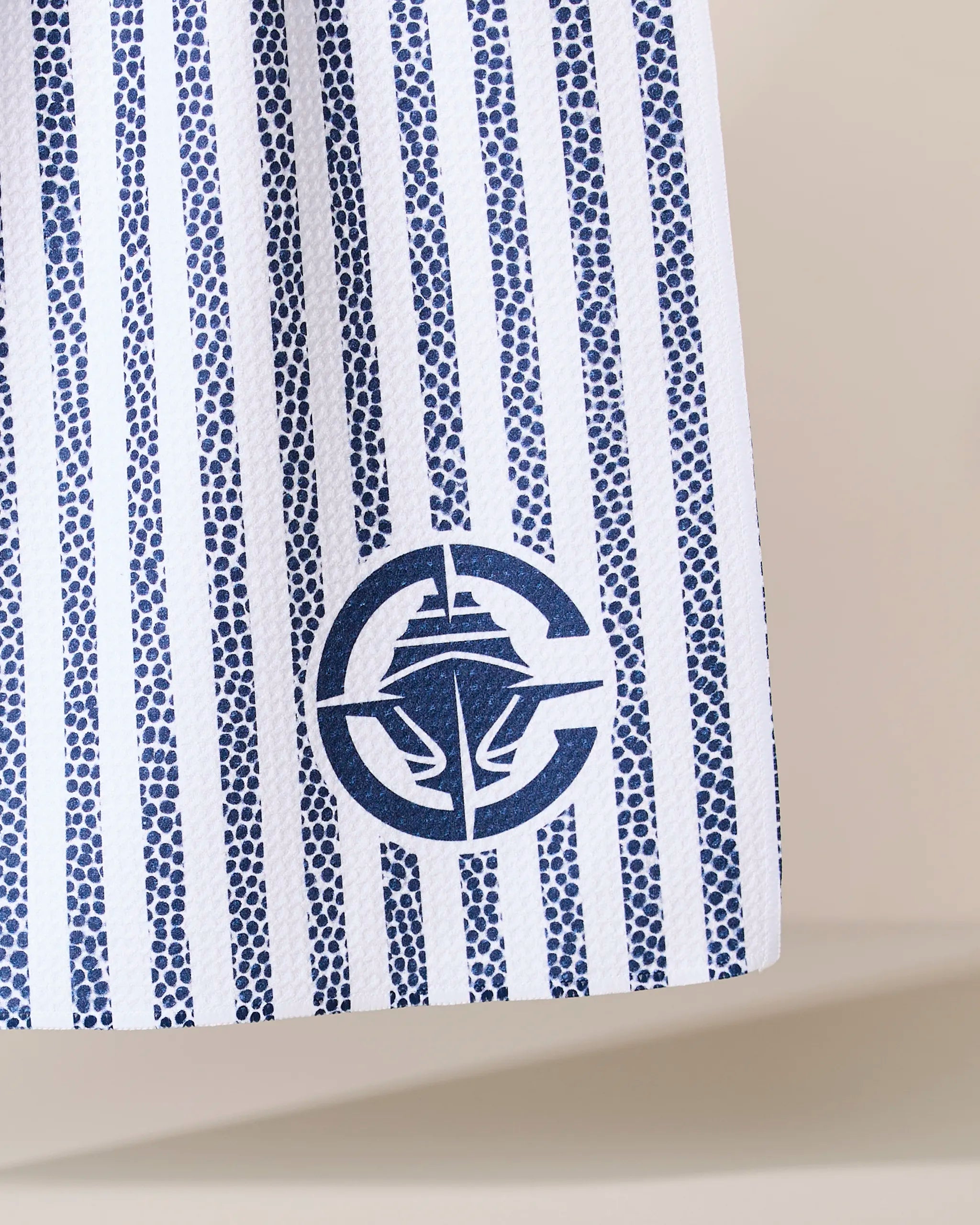 A close-up of Geometry’s Clippers Grip Stripe shows white fabric with vertical blue dotted stripes and a circular nautical logo—featuring a ship and partial anchor—near the hem.