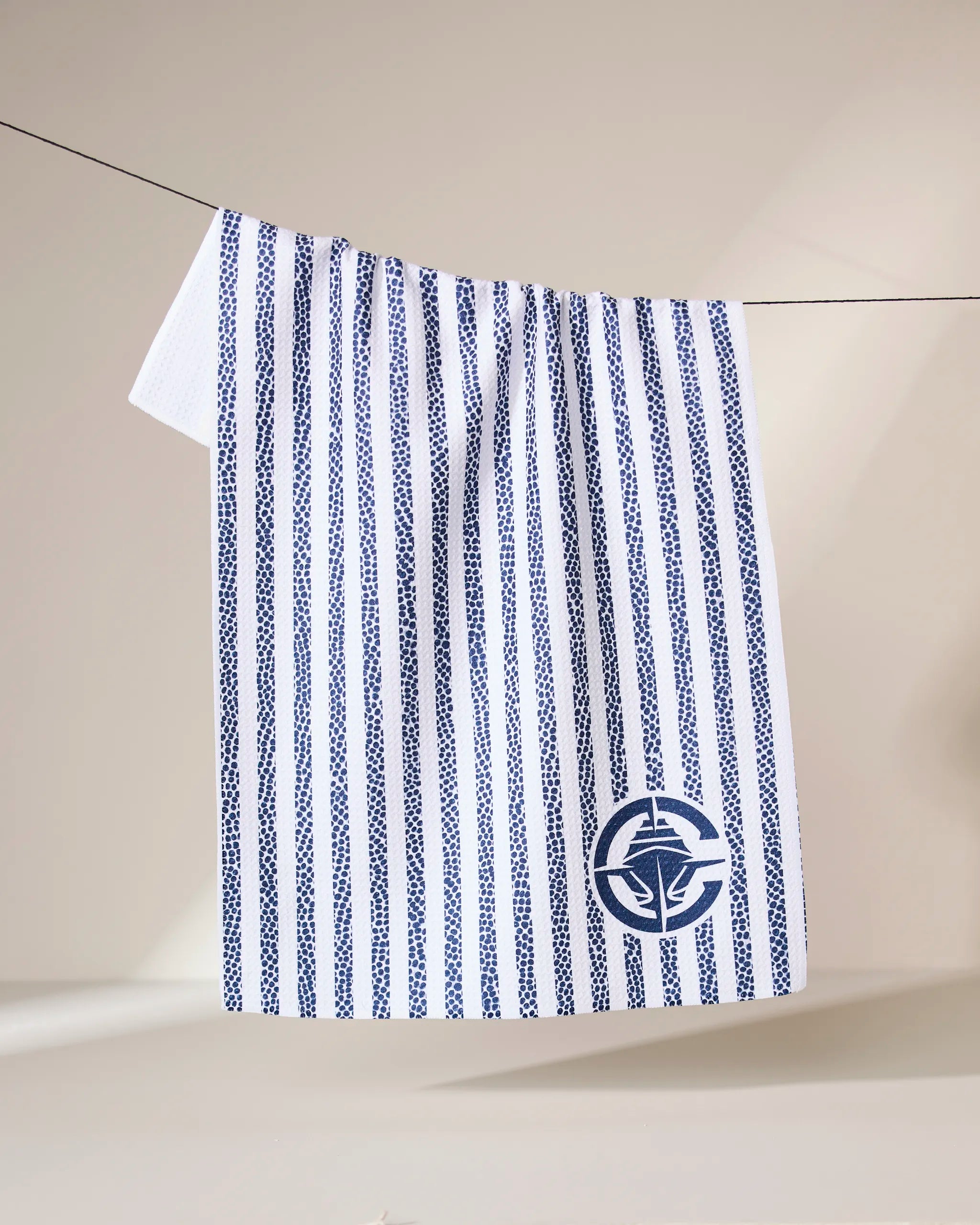 The Geometry Clippers Grip Stripe towel, featuring vertical navy blue stripes and a circular nautical emblem, hangs on a line against a neutral background.