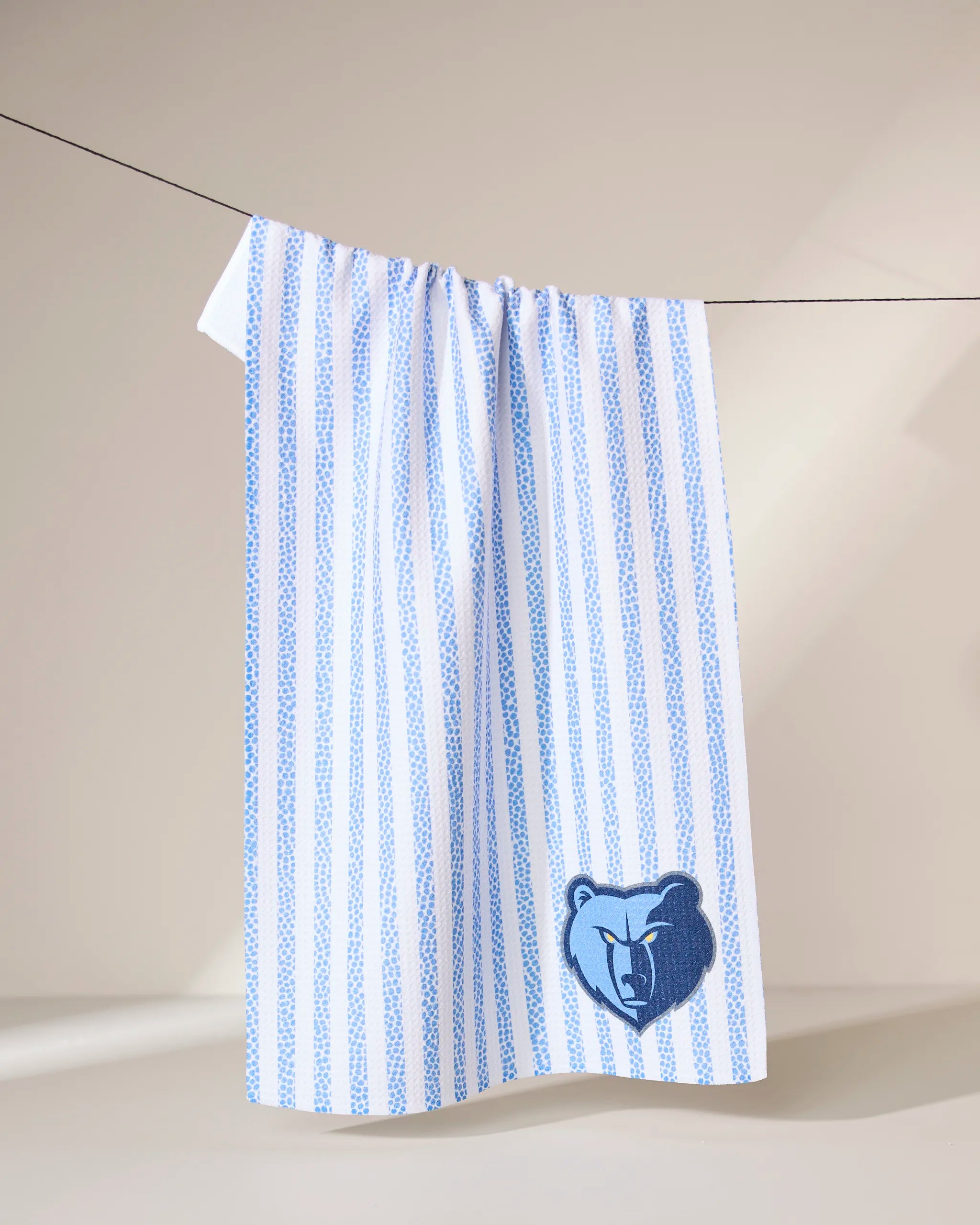 The Geometry Grizzlies Grip Stripe towel, featuring light blue vertical stripes and a dark blue embroidered bear head logo in the lower right corner, hangs on a clothesline against a softly lit neutral background.