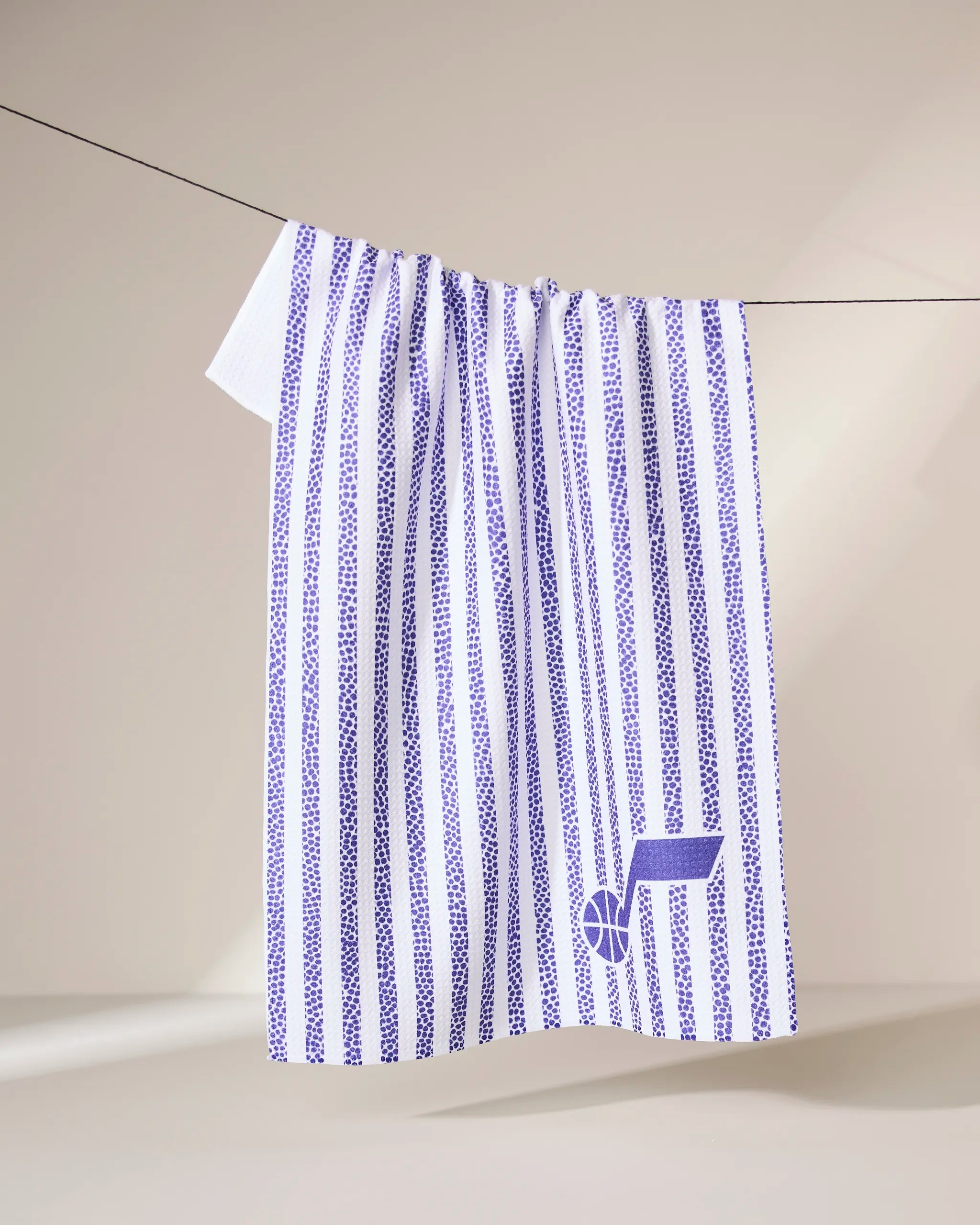 The Geometry Jazz Grip Stripe towel, featuring vertical blue dotted stripes and a small blue basketball logo, hangs neatly over a black clothesline against a neutral beige background.