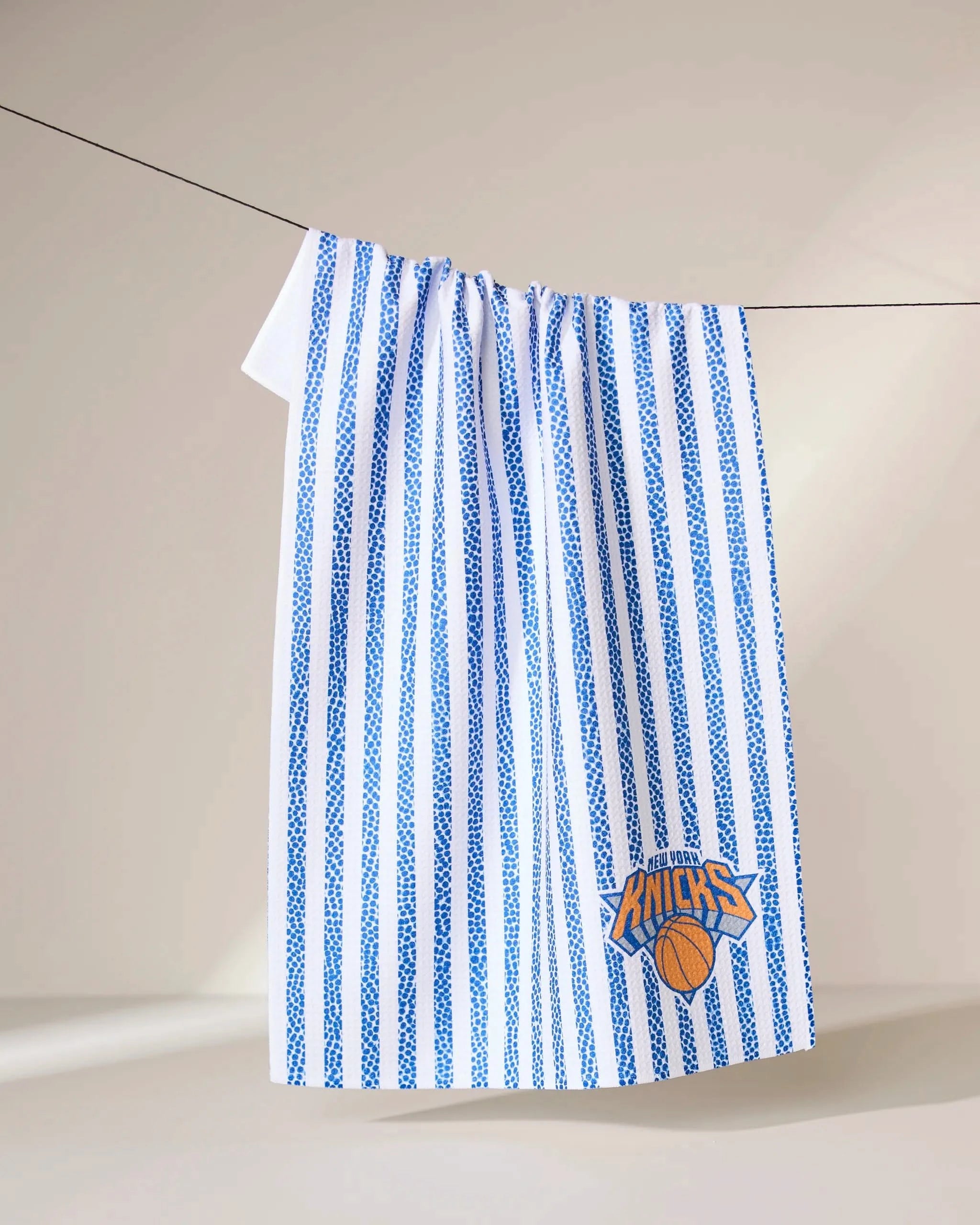 The Geometry Knicks Grip Stripe towel, featuring vertical blue stripes and a New York Knicks logo, hangs on a clothesline against a plain background.