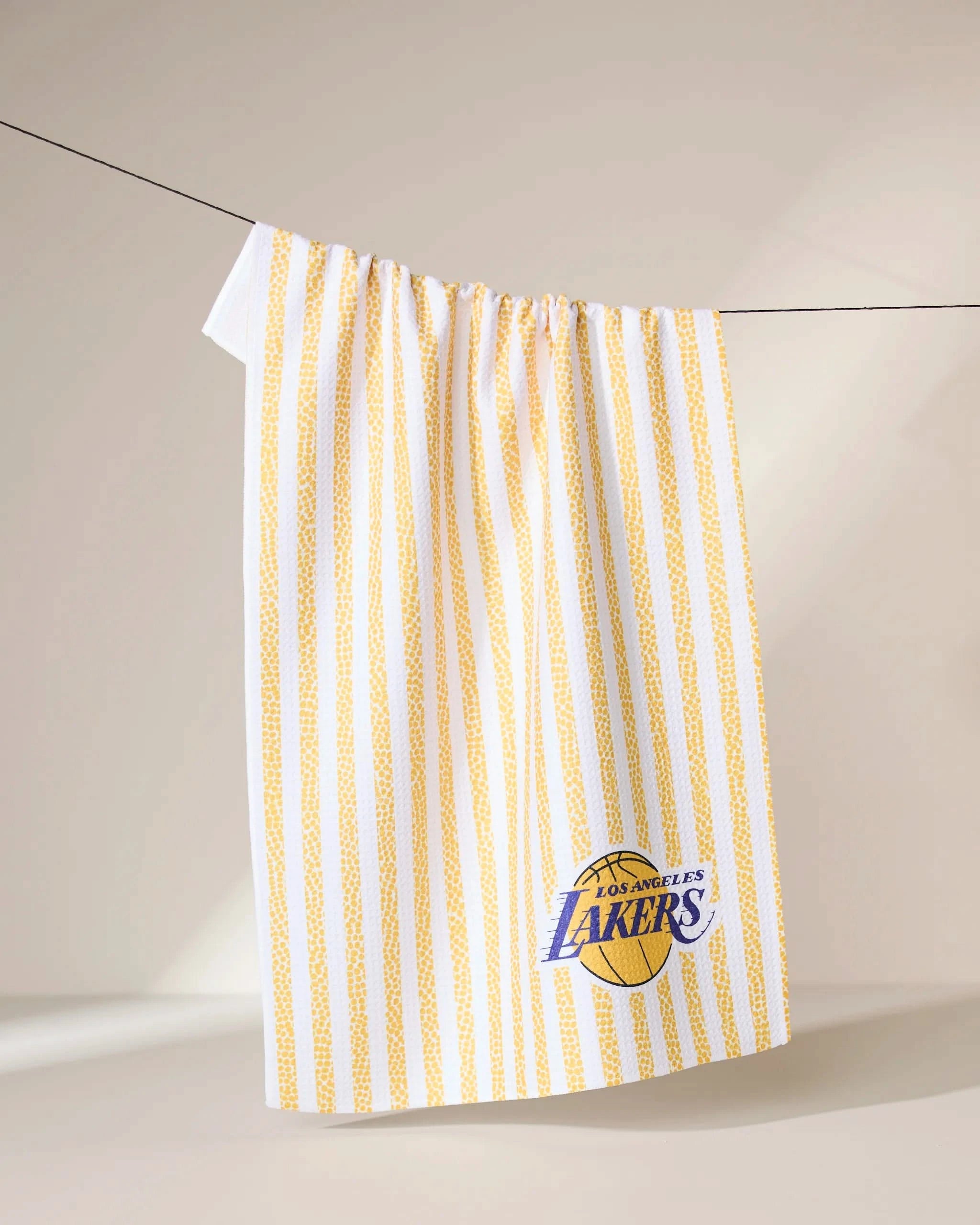 The Geometry Lakers Grip Stripe towel, featuring yellow vertical stripes and the Los Angeles Lakers logo, hangs over a black clothesline against a neutral background.