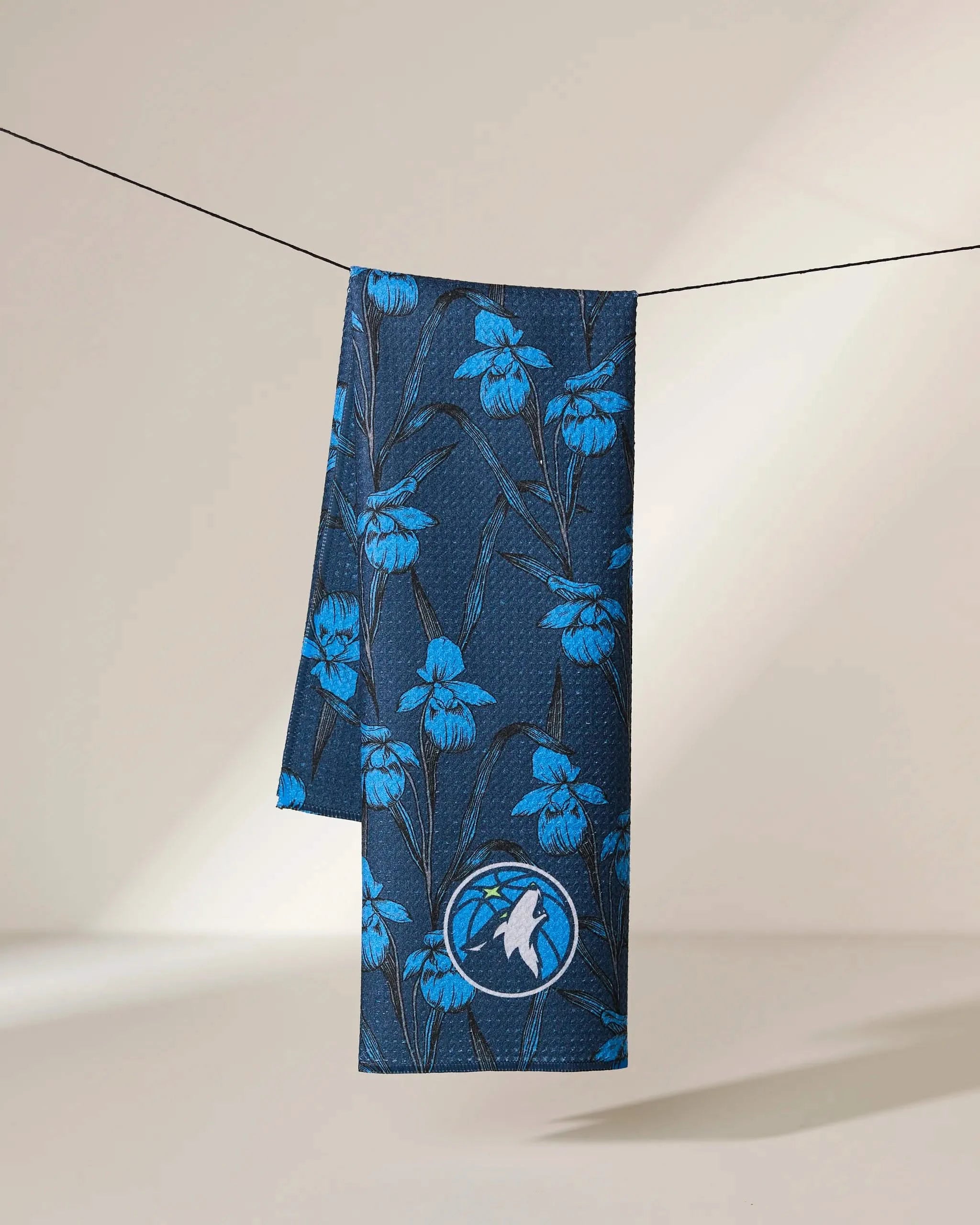 The Geometry Timberwolves Floral towel features a dark blue fabric with a bright blue floral pattern and a white bird patch, hanging on a black line against a softly lit beige background.
