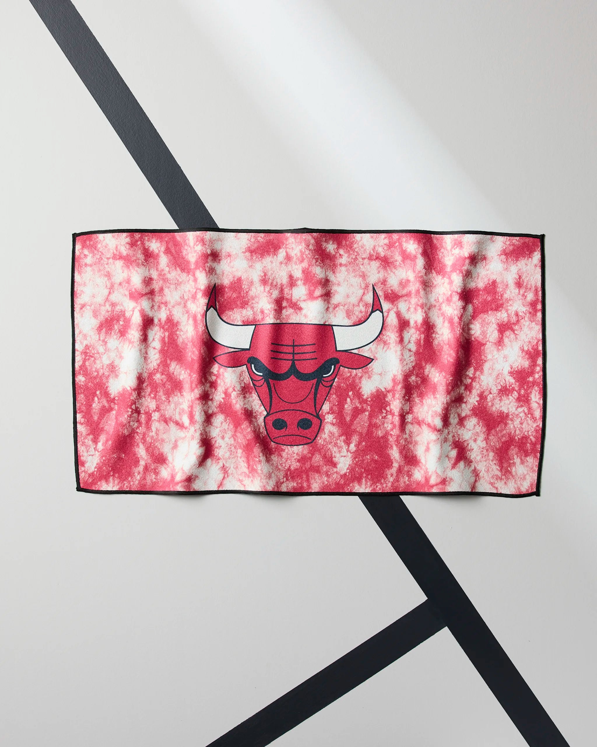 The Geometry Bulls Flash towel features a rectangular shape with a red and white tie-dye pattern and a cartoon bulls head in the center, displayed against a white wall with black diagonal lines.