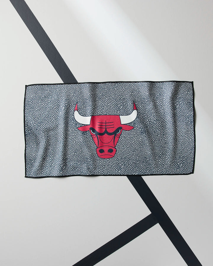 The NBALAB x Geometry Bulls Grip is a rectangular, quick-drying gym towel with a textured black-and-white pattern and a large red bull head logo with white horns outlined in black. It is shown laid flat on a light surface with intersecting black lines.