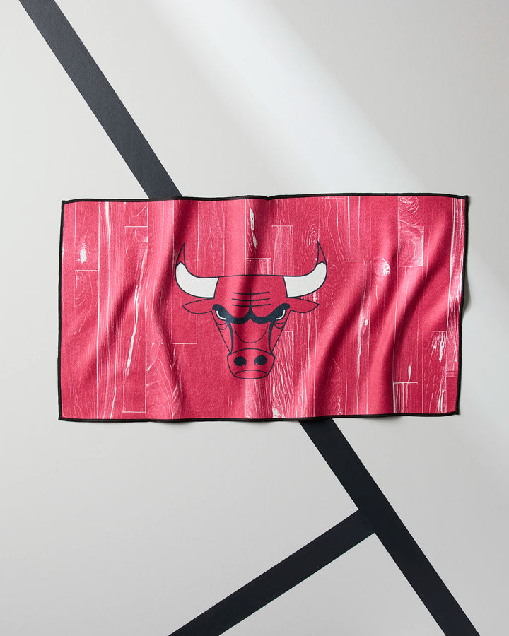 The Bulls Zone towel by NBALAB x Geometry, in red with a white-horned bull head logo, is quick-drying and displayed on a light gray surface accented by diagonal black lines.
