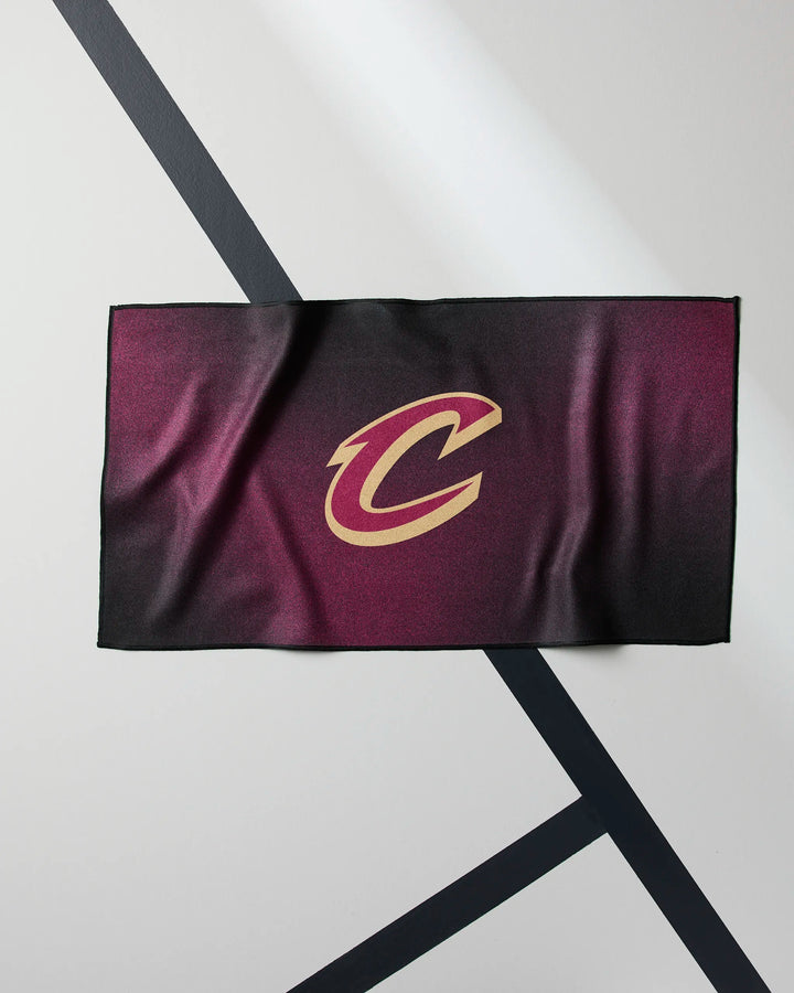 The Cavaliers Fade fitness towel by NBALAB x Geometry, featuring a gold C logo in maroon and black, is displayed flat on a light surface with intersecting black diagonal lines.