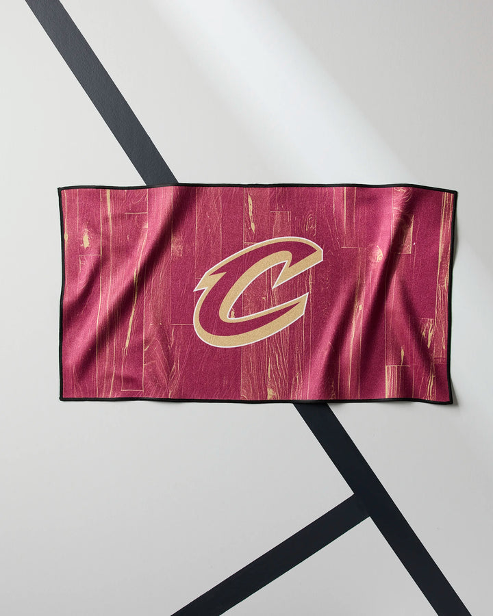 The Cavaliers Zone fitness towel by NBALAB x Geometry features a maroon design with a prominent gold C logo, inspired by the Cleveland Cavaliers, and is made from high-performance Geoweave fabric. Displayed on a white wall with black diagonal lines.
