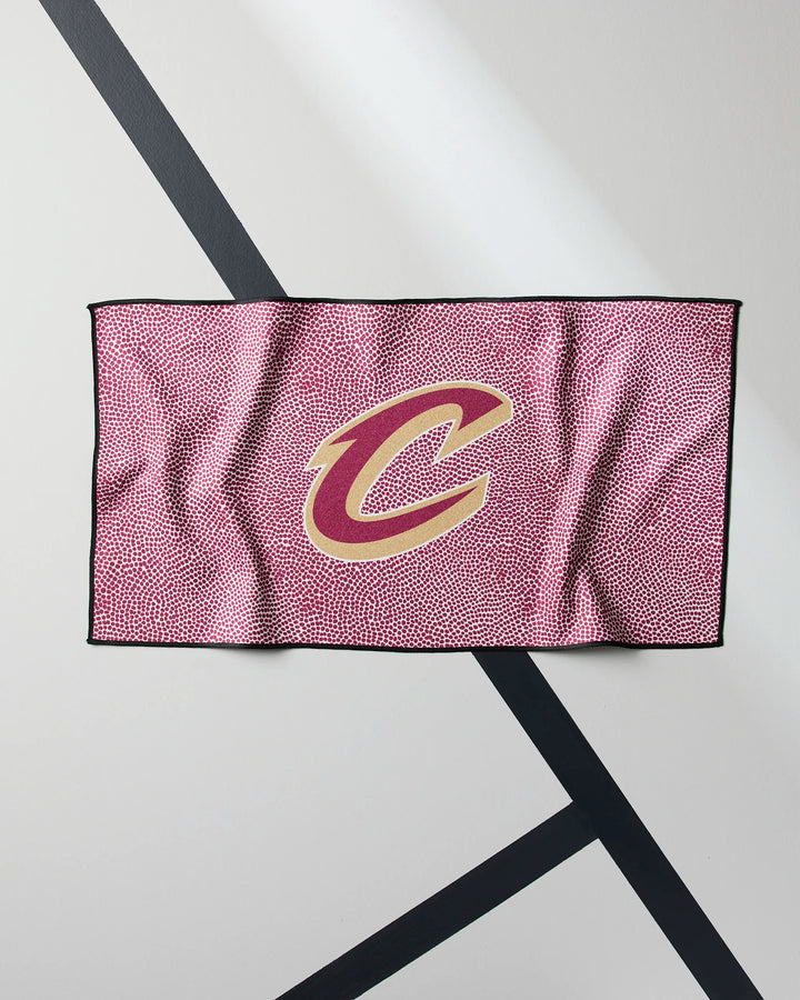 The NBALAB x Geometry Cavaliers Grip is a rectangular fitness towel featuring a repeating basketball texture and a bold gold and red Cleveland Cavaliers C logo in the center, shown hanging on a white wall with black diagonal stripes.