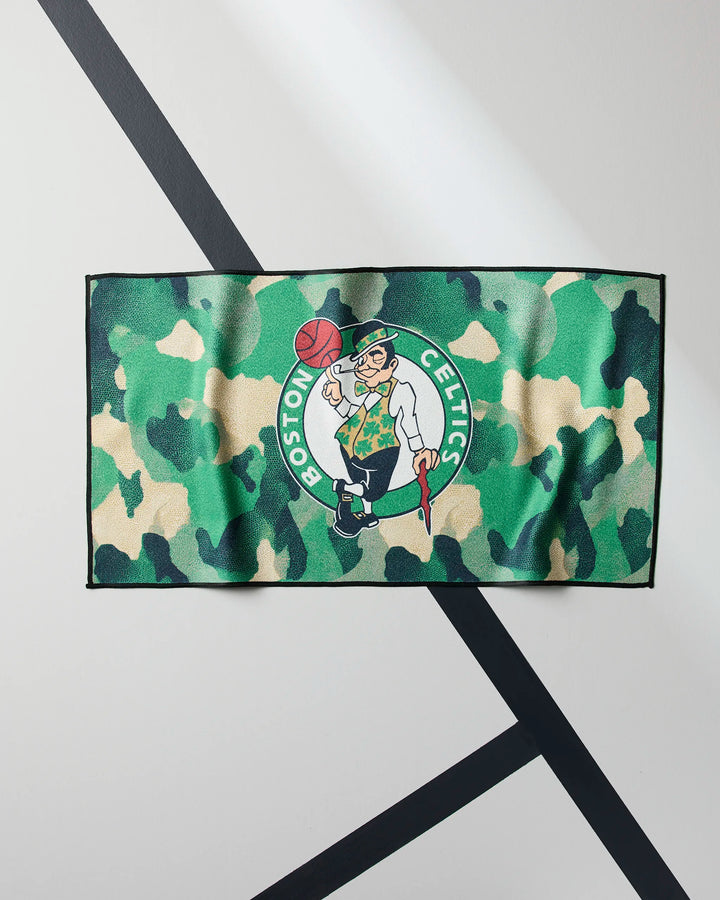 The Celtics Camo by NBALAB x Geometry is a rectangular fitness towel with a green camo pattern and Boston Celtics logo, pictured on a white floor with black intersecting stripes.