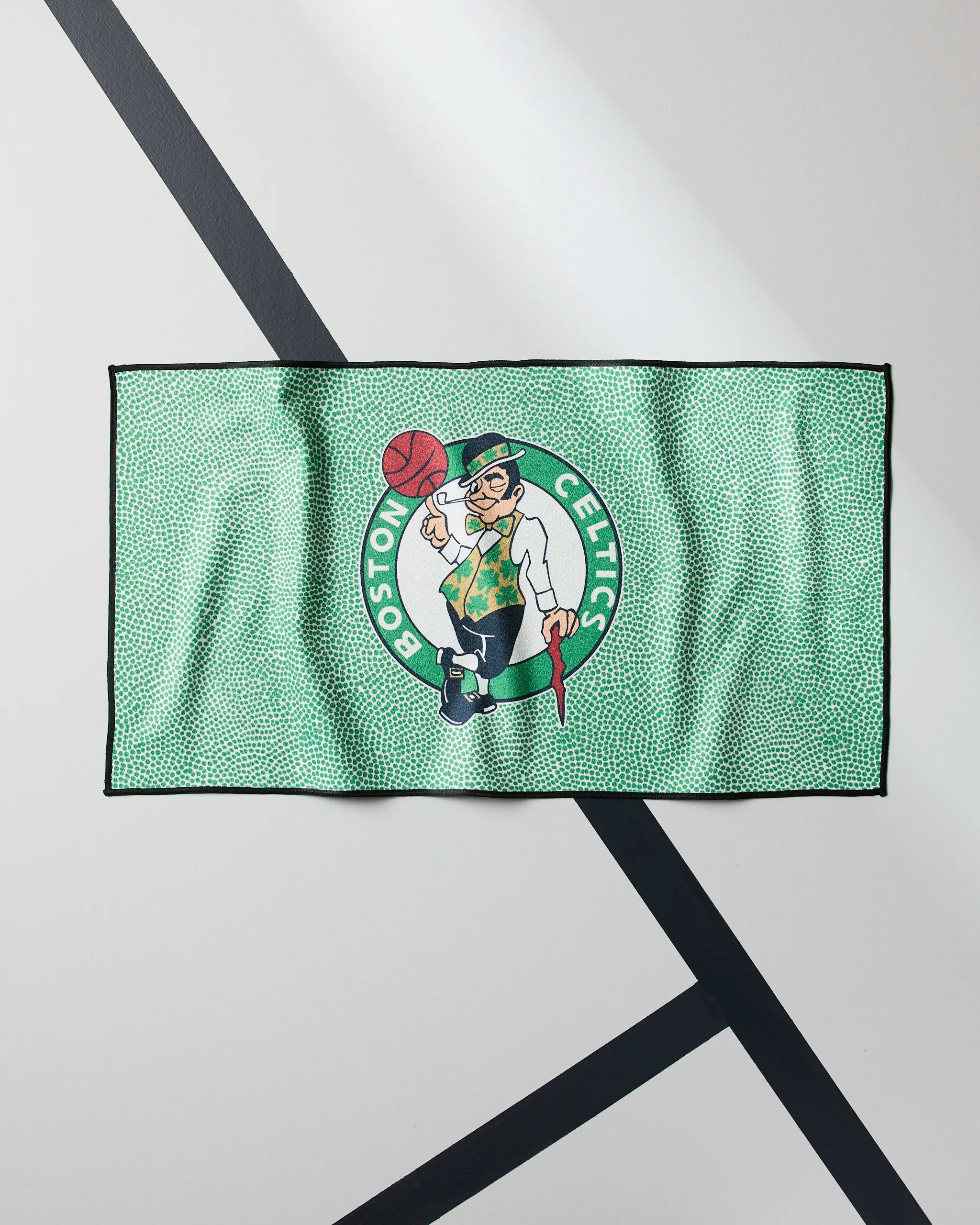 The Geometry Celtics Grip is a green NBA fitness towel featuring the Boston Celtics logo and leprechaun mascot spinning a basketball, shown flat on a white surface with black diagonal lines underneath.