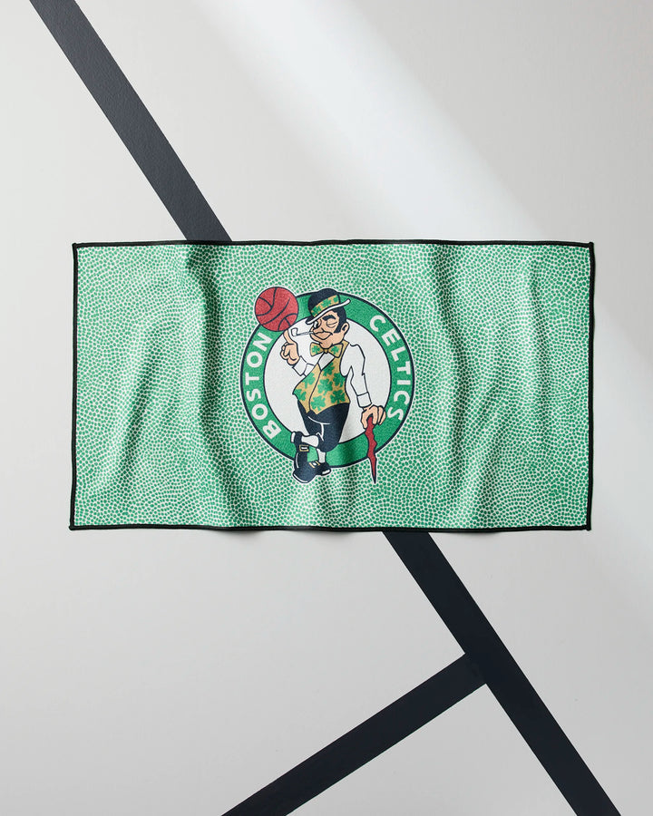 The Celtics Grip by NBALAB x Geometry is a green fitness towel featuring the Boston Celtics logo with a leprechaun spinning a basketball, shown laid flat on a light surface with black intersecting lines.