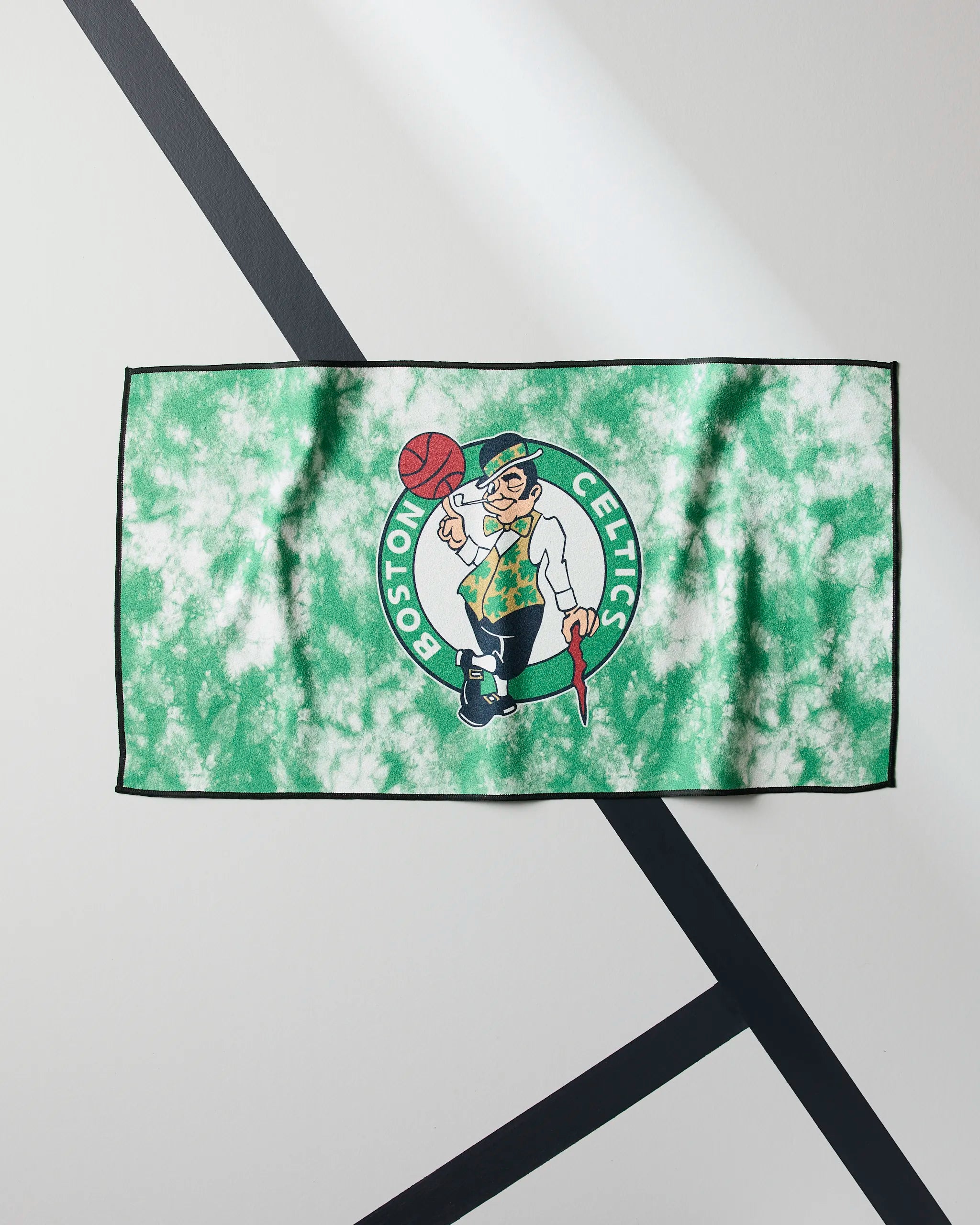 The Celtics Flash by Geometry is a green and white tie-dye NBA fitness towel featuring the Boston Celtics logo with a leprechaun spinning a basketball, displayed on a surface with intersecting black lines.