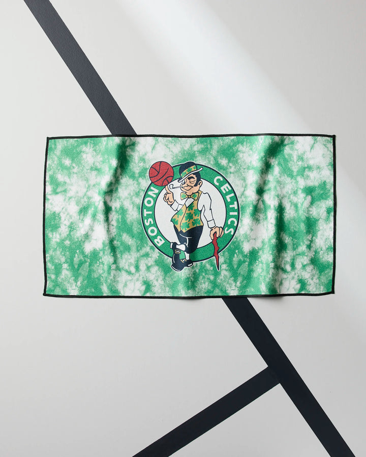 The NBALAB x Geometry Celtics Flash towel, in green and white tie-dye with the Boston Celtics leprechaun logo, is laid flat on a floor marked with black intersecting lines.