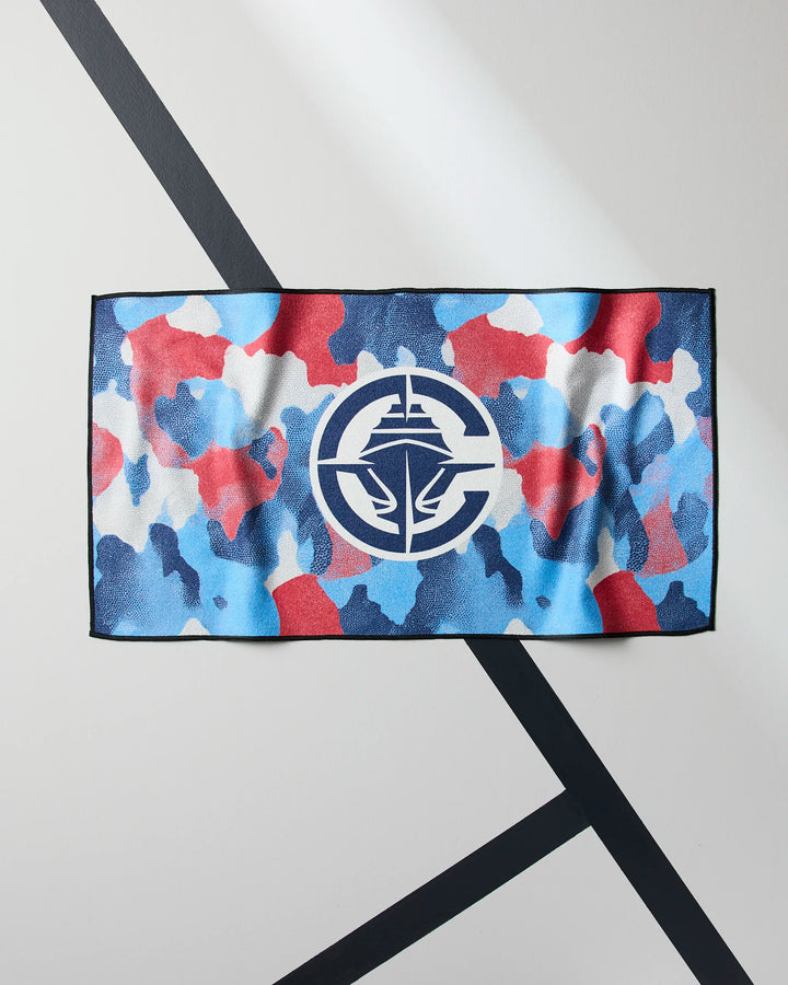 The Clippers Camo gym towel by NBALAB x Geometry is rectangular, quick-drying, and features a red, blue, and white camouflage pattern with a central white ship emblem. It’s displayed on a white background with dark diagonal lines.