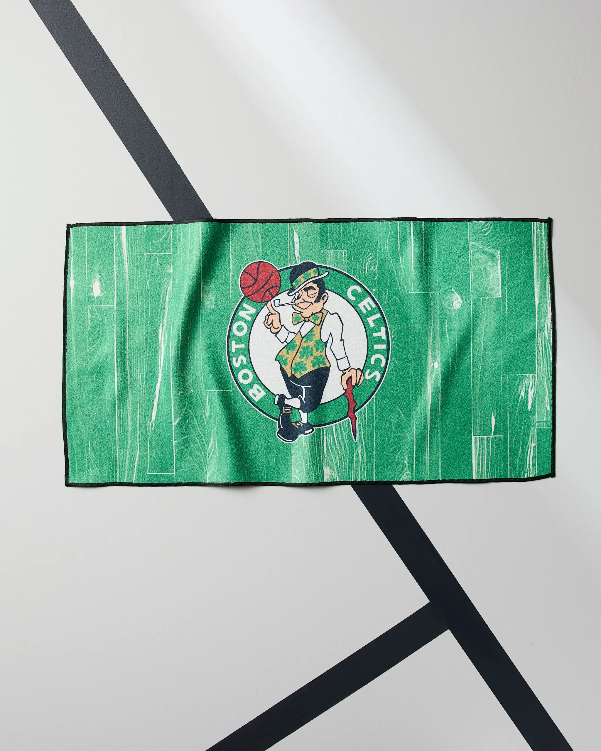 The Celtics Zone fitness towel by Geometry, featuring the Boston Celtics leprechaun logo in green, is displayed on a pale surface with intersecting black lines.